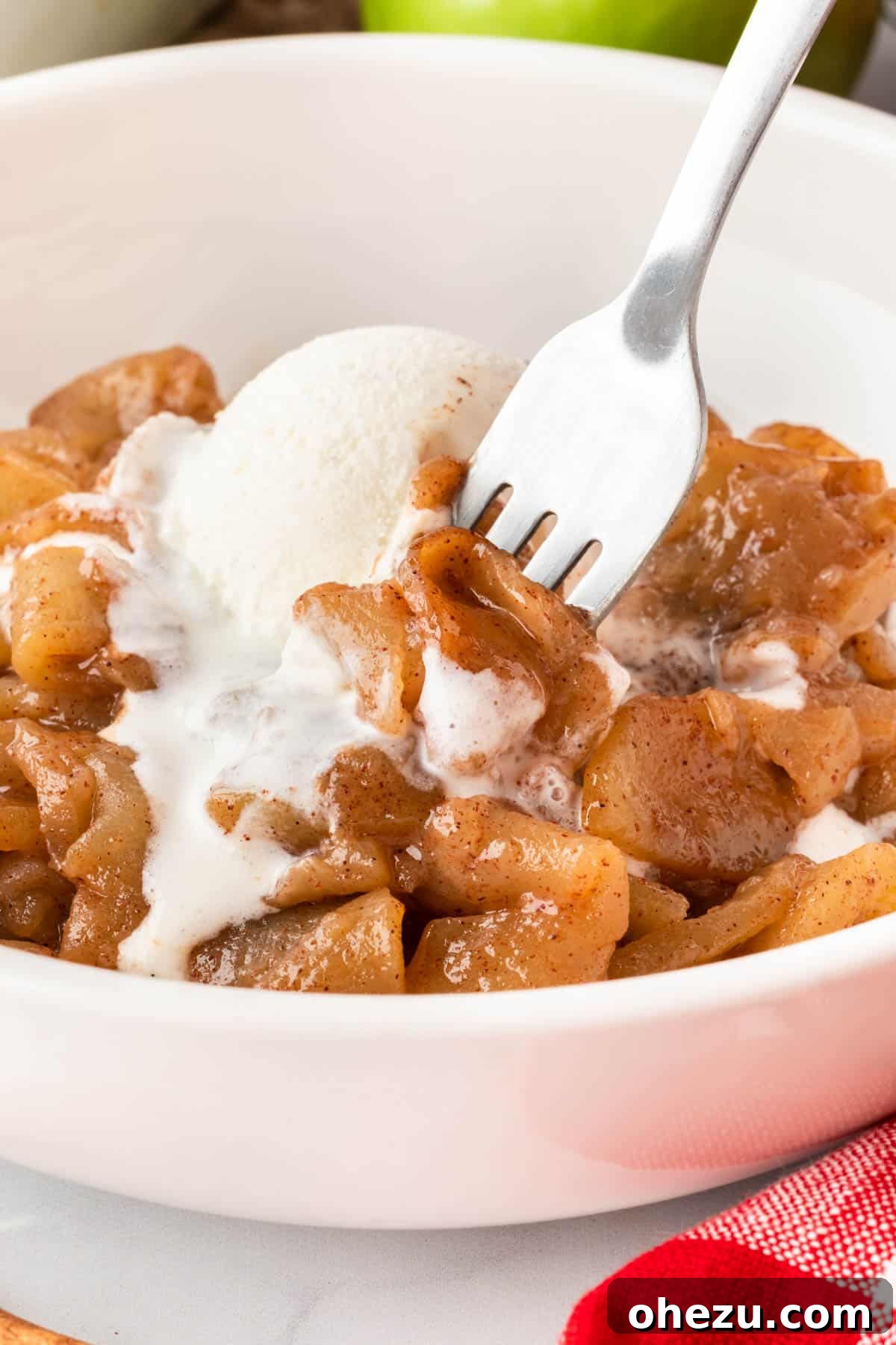 A fork taking a bite of baked cinnamon apples, showcasing the tender apple slice, rich caramelized sauce, and a hint of melting vanilla ice cream.