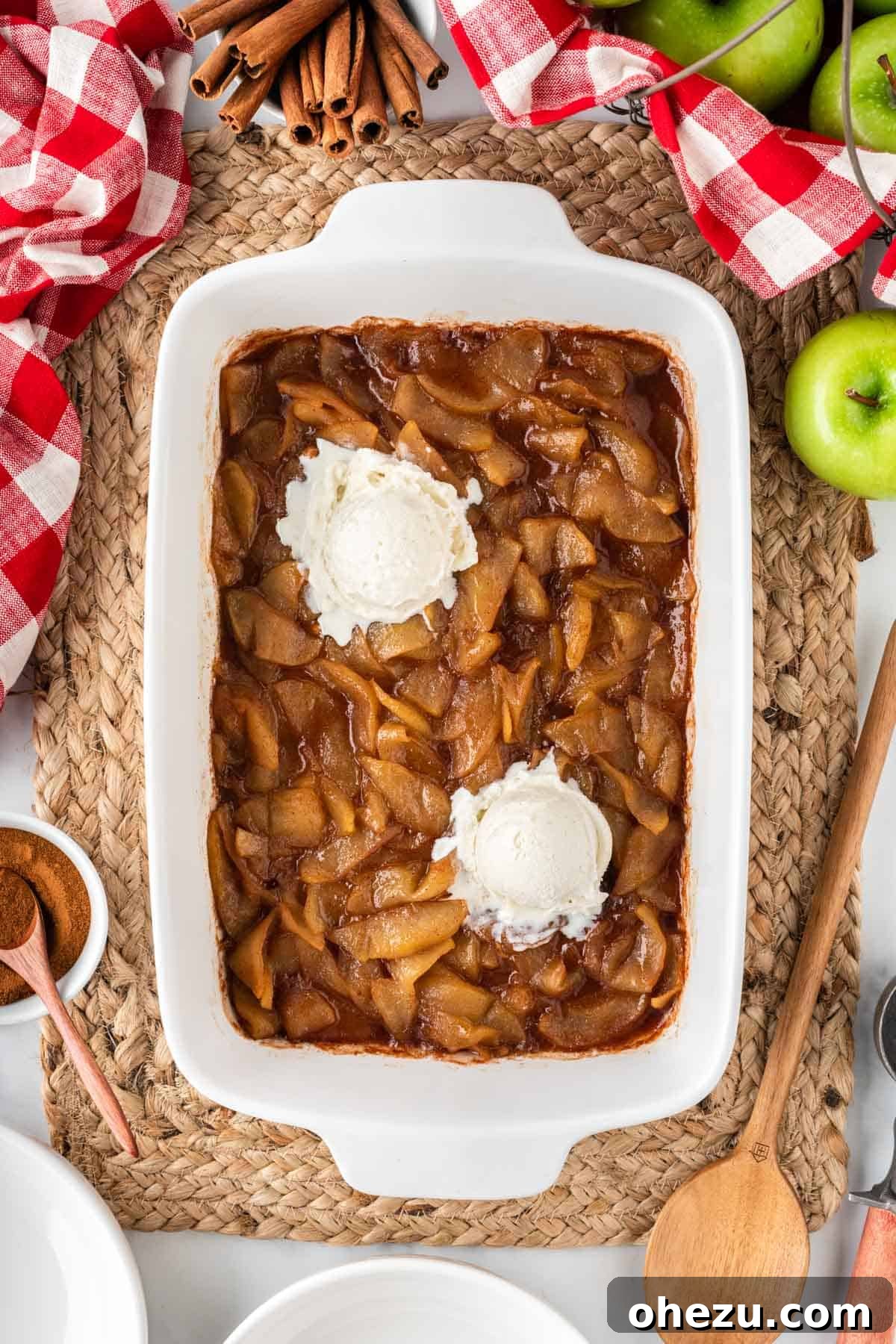 A wide shot of baked cinnamon apples in a white rectangular baking dish, generously topped with multiple scoops of melting vanilla ice cream, creating a mouth-watering fall dessert spread.