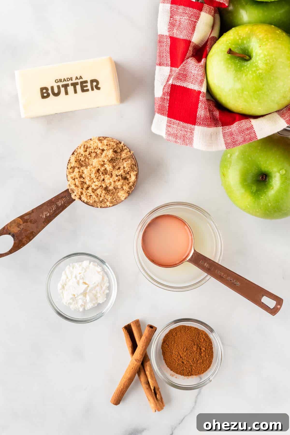 A flat lay photograph showcasing all the simple ingredients needed to make baked cinnamon apples: fresh Granny Smith apples, brown sugar, cornstarch, butter, cinnamon, and a lemon.