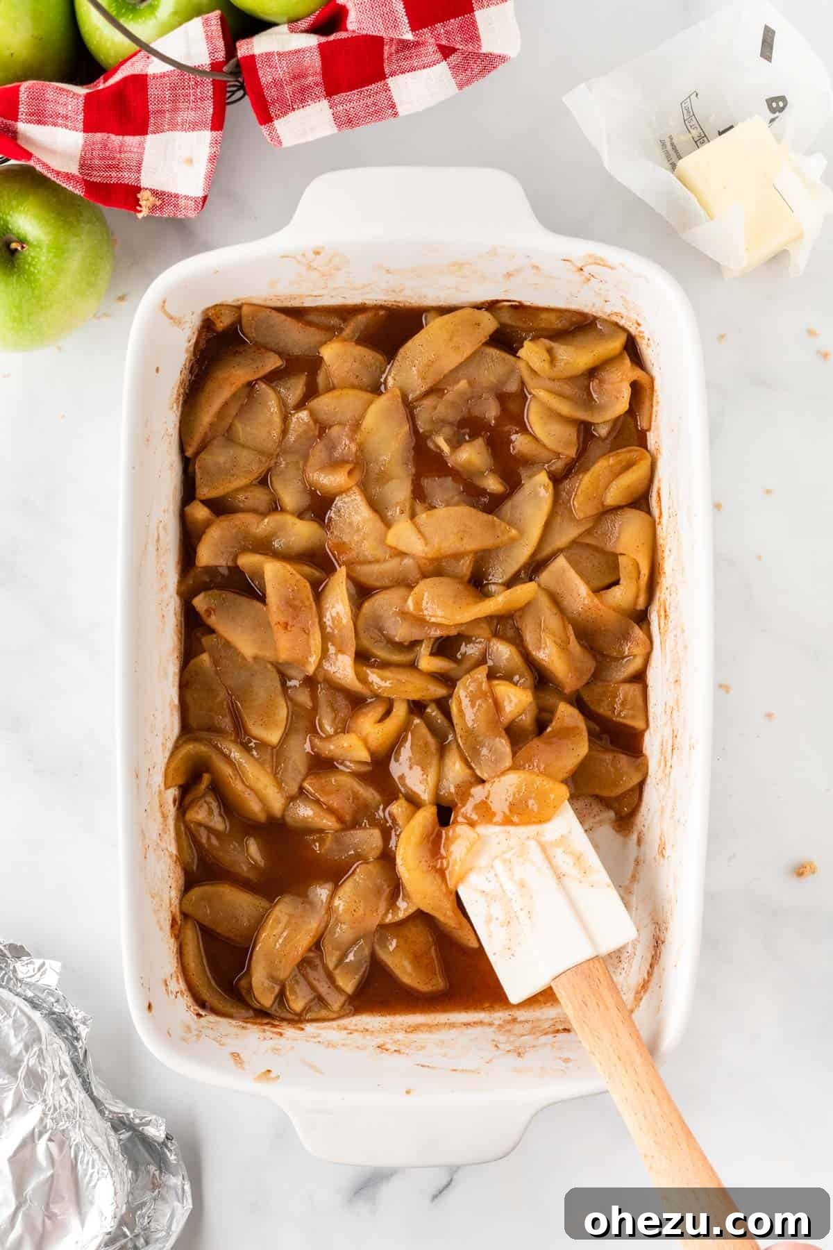 A wooden spoon stirring the baked cinnamon apples in the baking dish, showing the bubbly, golden-brown caramelized sauce and softened apple slices.