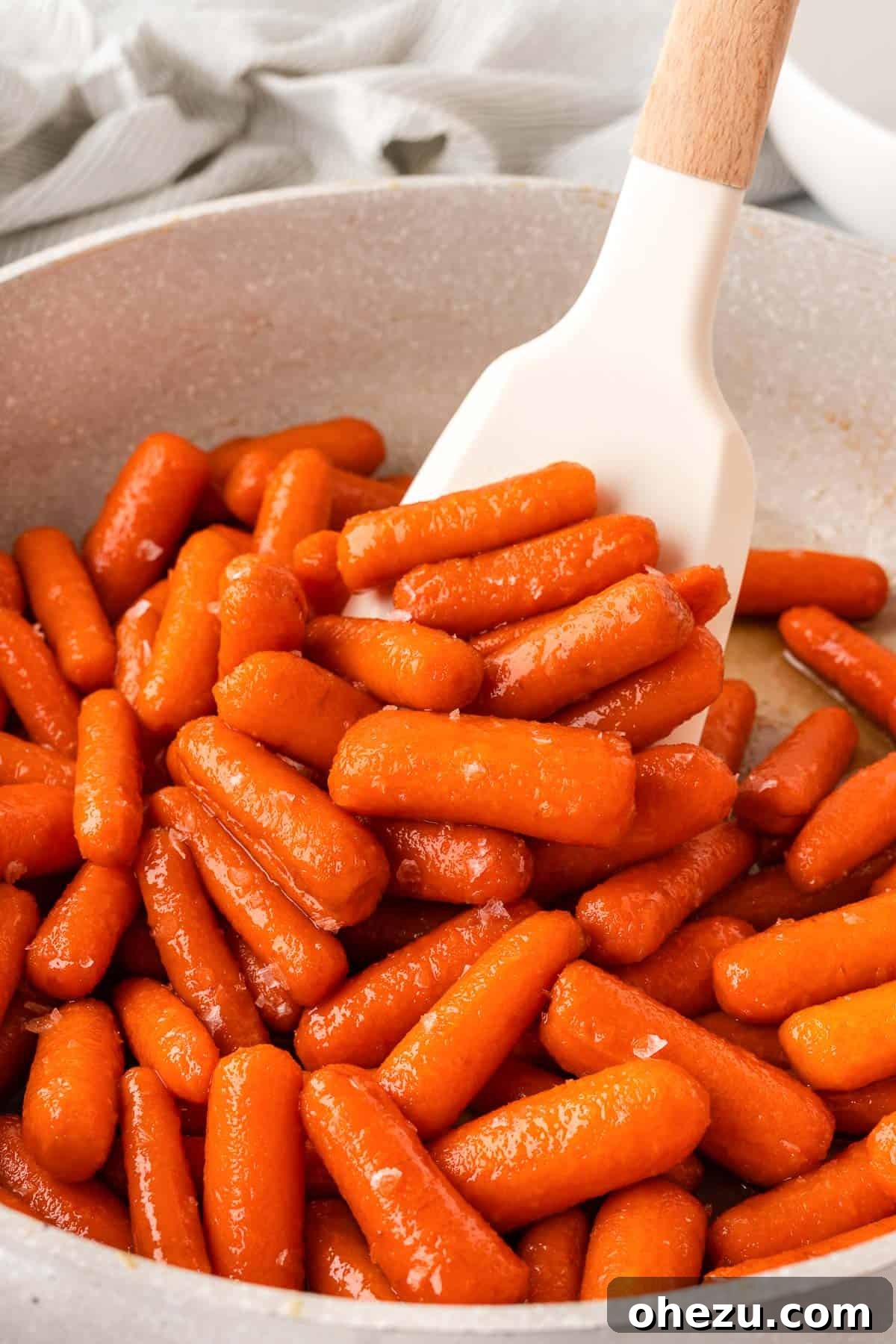A close-up view of vibrant orange baby carrots being gently stirred in a skillet, coated in a rich, bubbling brown sugar and honey glaze.