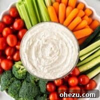 A vibrant bowl of creamy veggie dip surrounded by an array of colorful, fresh raw vegetables including broccoli florets, bright red cherry tomatoes, crisp celery sticks, and sweet baby carrots.