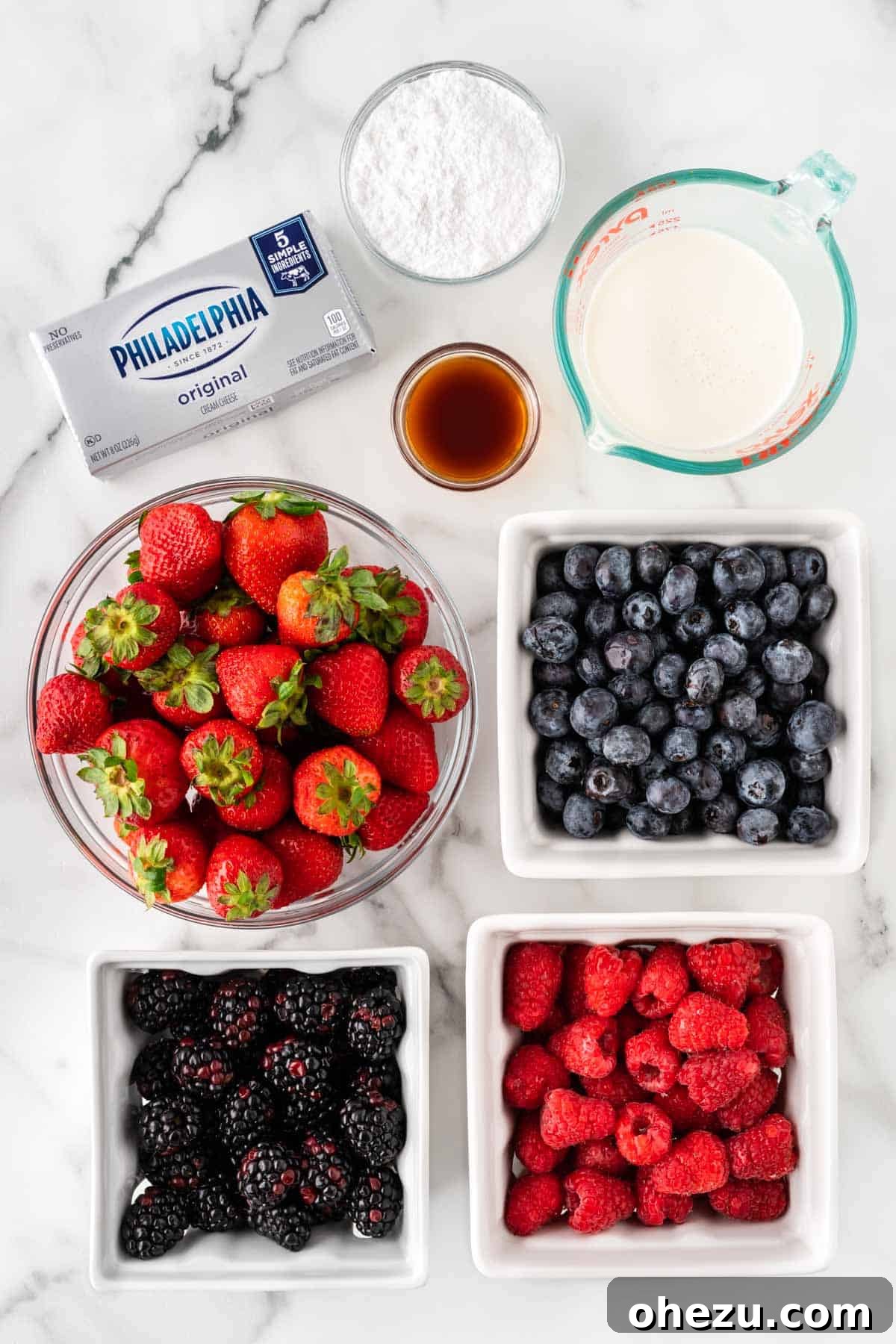 A collection of ingredients laid out on a table, including heavy cream, cream cheese, powdered sugar, vanilla extract, and a variety of fresh berries like strawberries, blueberries, blackberries, and raspberries.