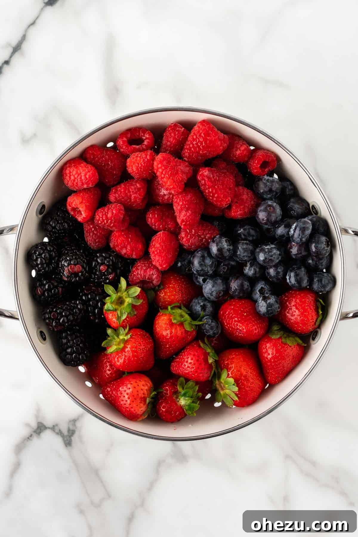 Freshly washed raspberries, blackberries, blueberries, and sliced strawberries resting in a colander, glistening with water droplets.