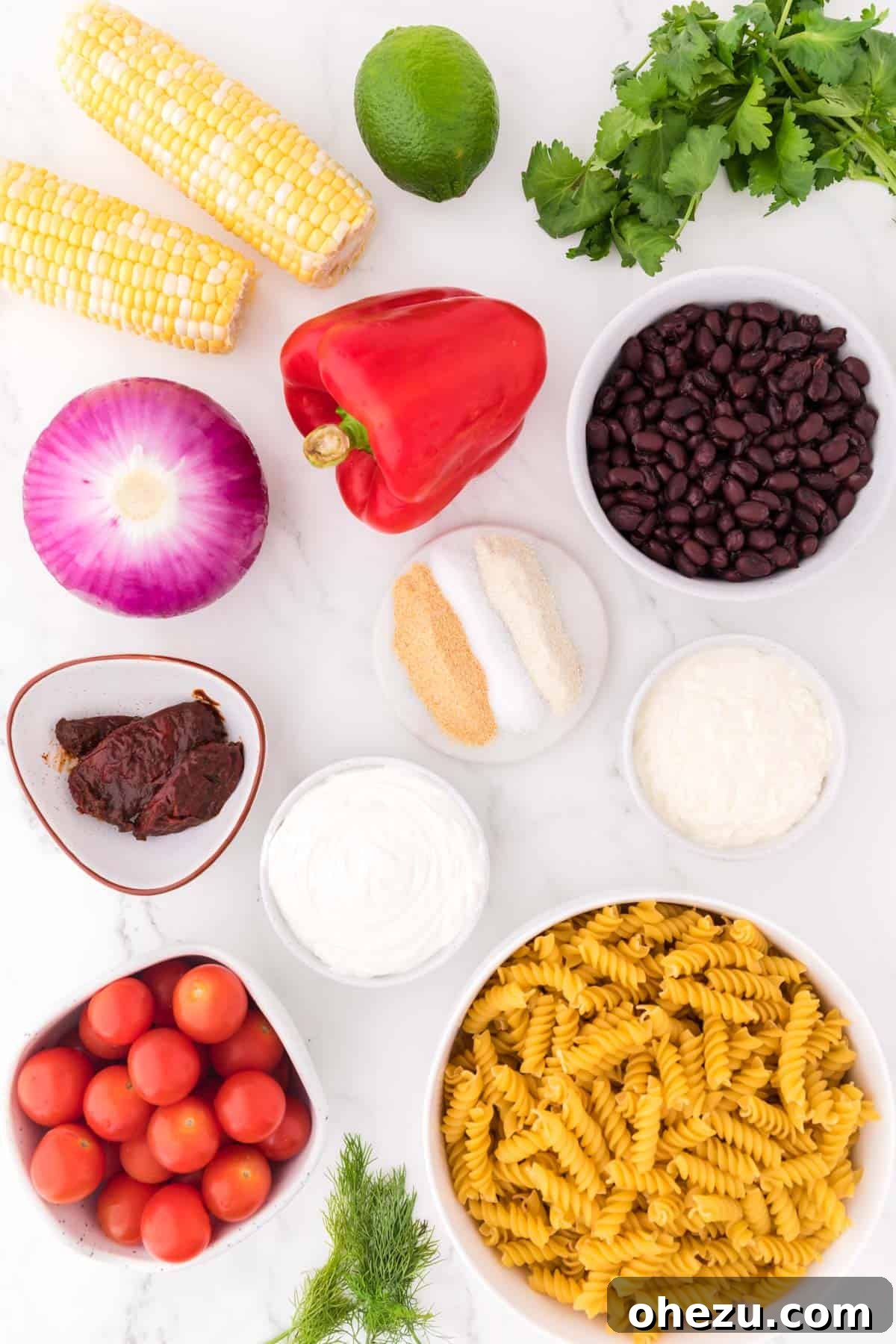 Zesty Fiesta Pasta Salad 3 All the fresh ingredients laid out on a white surface, including rotini pasta, black beans, corn on the cob, red bell pepper, red onion, cherry tomatoes, cilantro, and the ingredients for the chipotle ranch dressing.