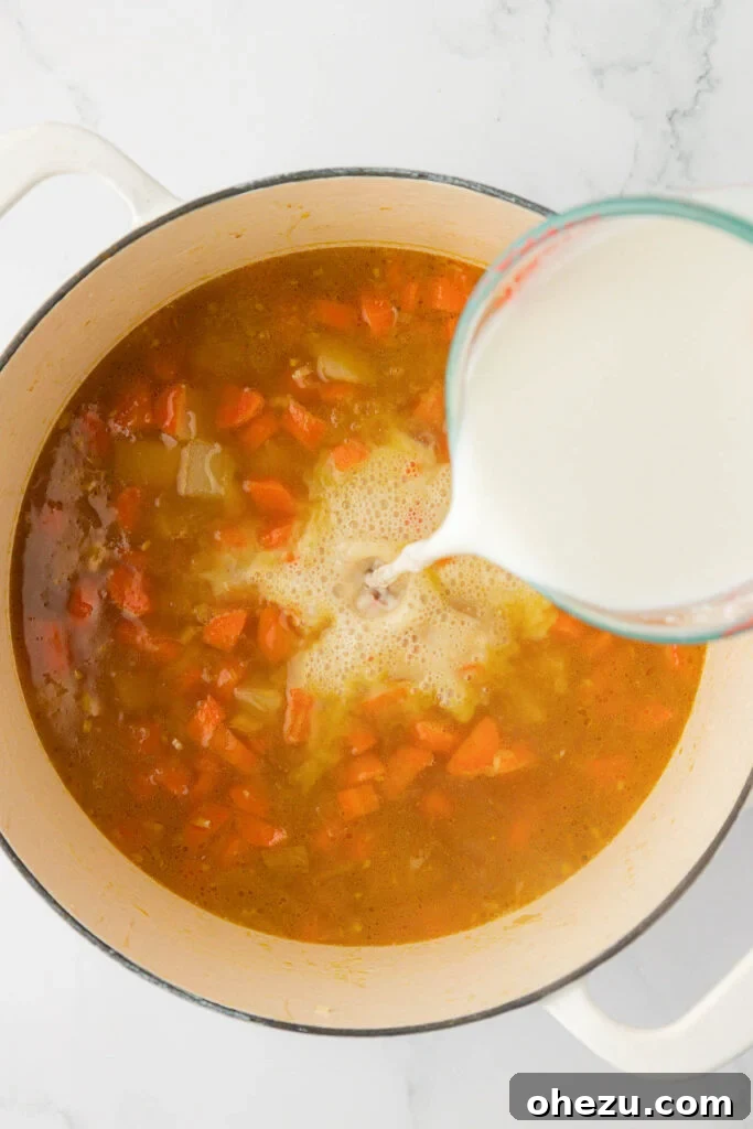 A steady, controlled stream of the milk and flour slurry being poured into the simmering vegetable soup, with a spoon nearby for constant stirring to ensure smooth incorporation.