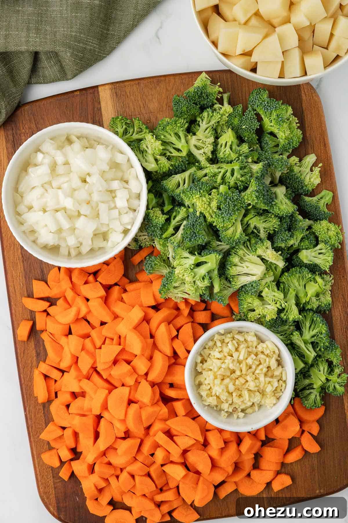 A wooden cutting board showcasing an array of meticulously chopped vegetables including finely diced carrots, onions, minced garlic, and perfectly portioned broccoli florets, all prepped and ready to be added to the creamy vegetable soup.
