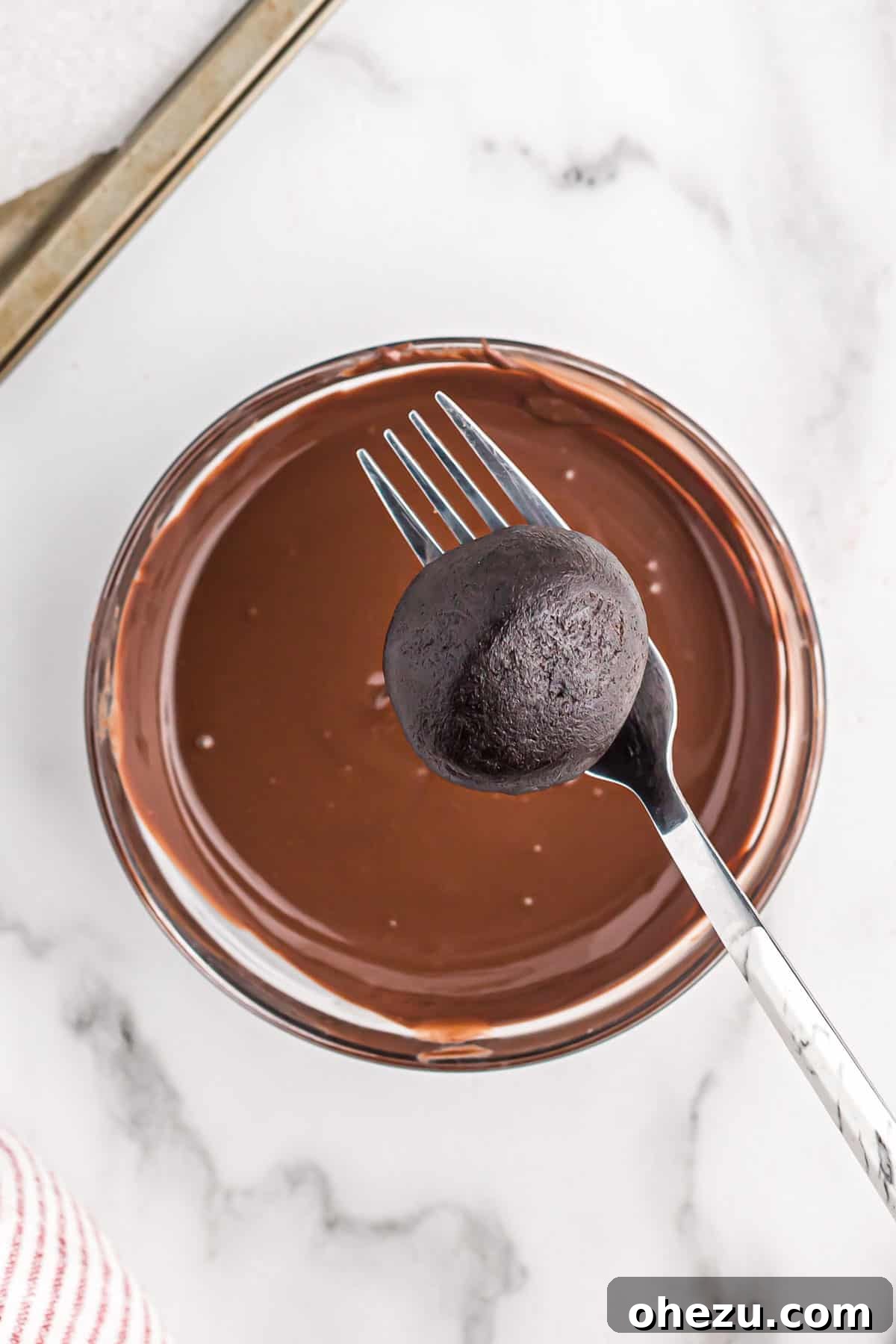 A hand holding an oreo ball on a fork, poised over a bowl of melted dark chocolate, ready for dipping.