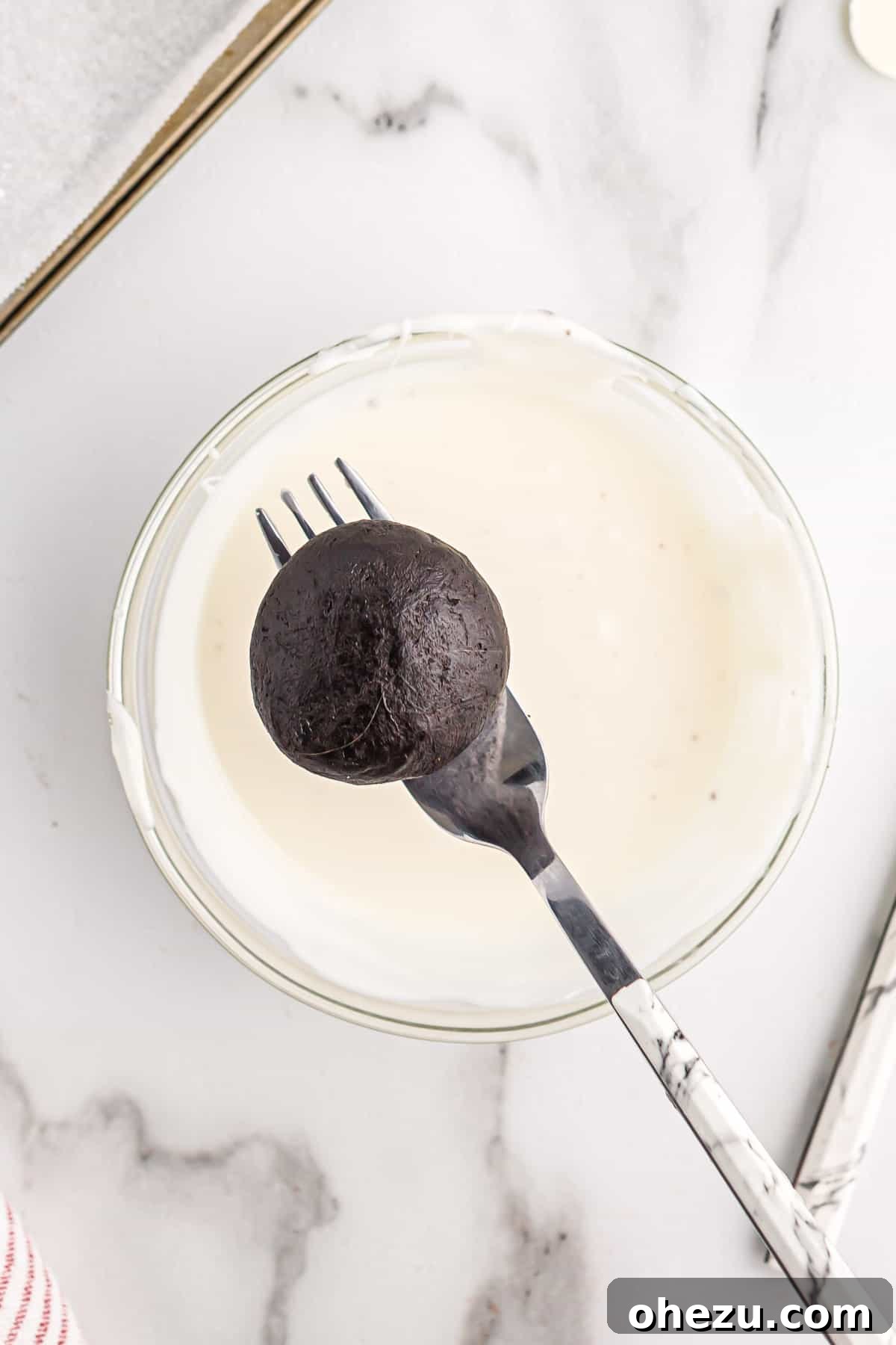 A hand holding an oreo ball on a fork, ready to be dipped into a bowl of creamy melted white chocolate.
