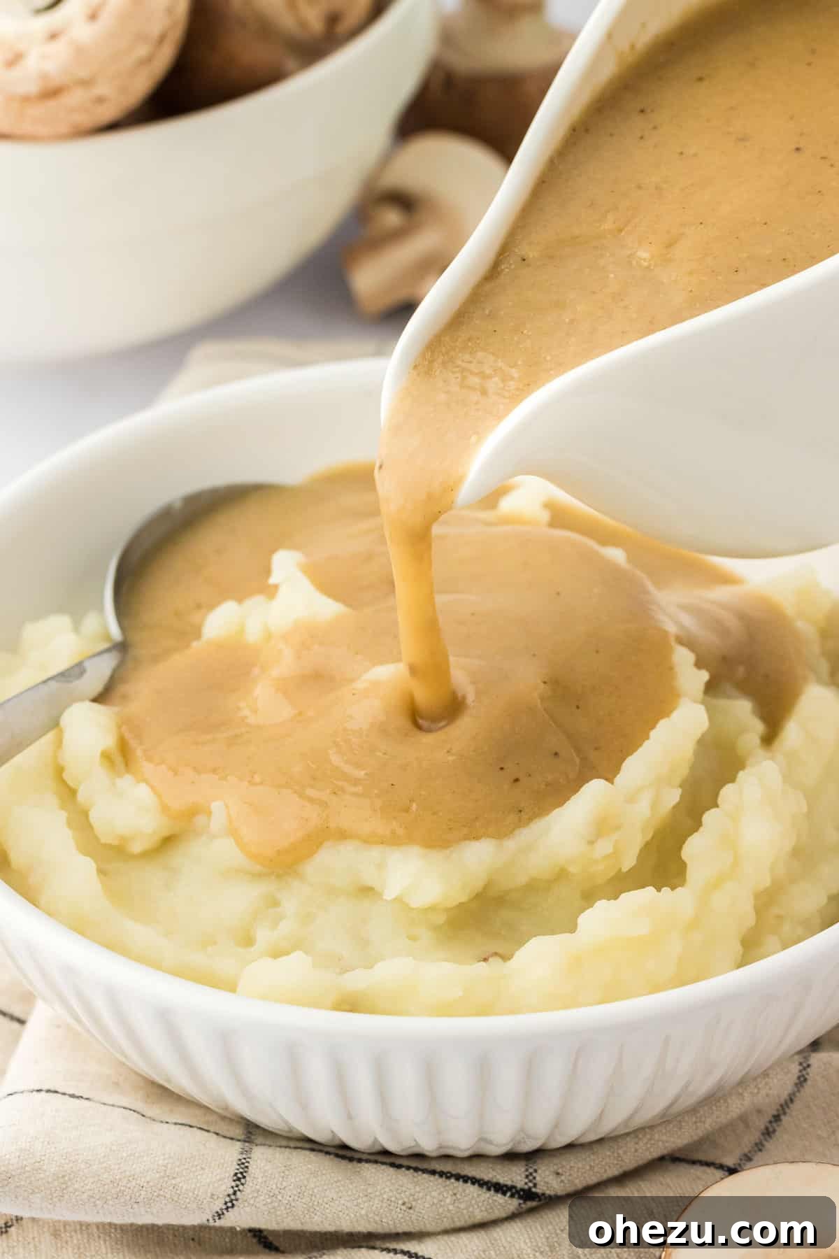 A hand pouring a rich, luscious stream of homemade vegetarian gravy from a pristine white gravy boat onto a serving of creamy mashed potatoes, ready to be devoured.