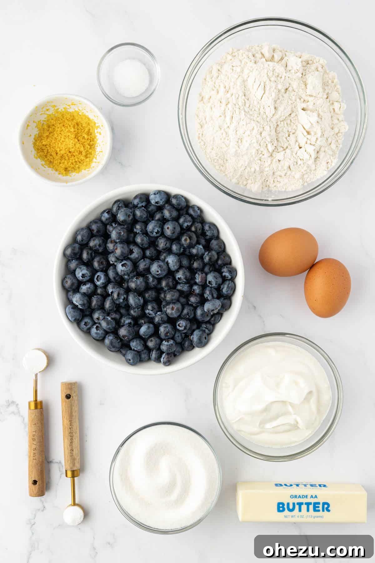 A selection of fresh ingredients laid out on a counter, including flour, sugar, eggs, butter, sour cream, lemon, and a bowl of fresh blueberries, ready for making muffins.