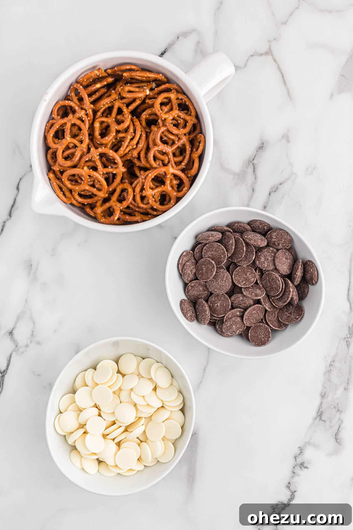 Three small bowls containing mini pretzels, dark chocolate melting wafers, and white vanilla melting wafers, illustrating the simple ingredients needed for this recipe.