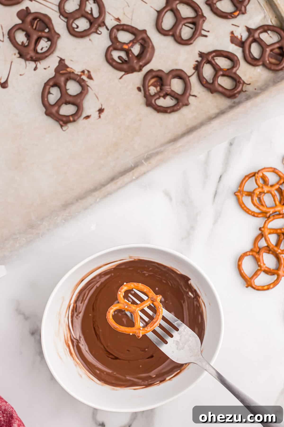 A hand holding a mini pretzel over a small bowl of melted dark chocolate, ready for dipping.