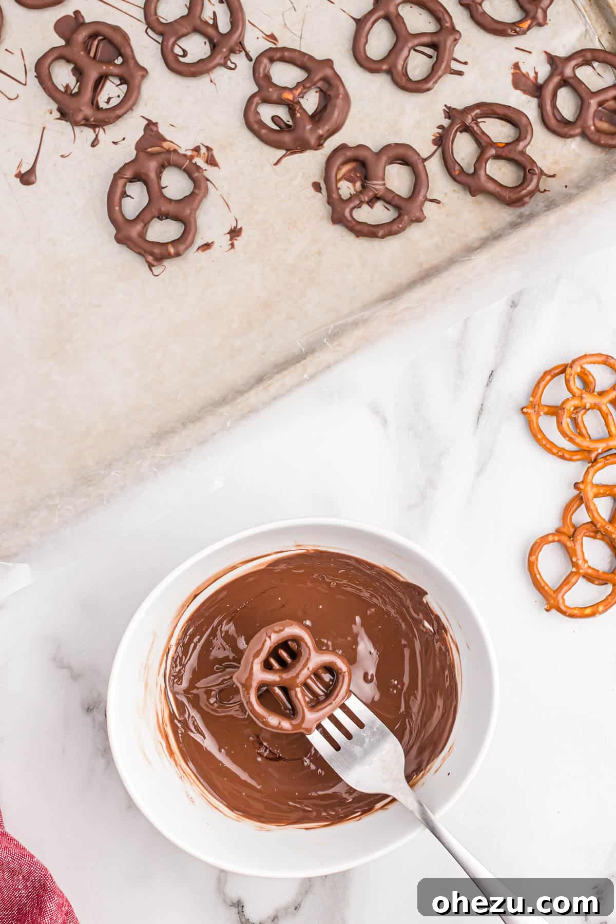 A mini pretzel fully dipped in melted dark chocolate, with excess chocolate dripping off, demonstrating the coating process.