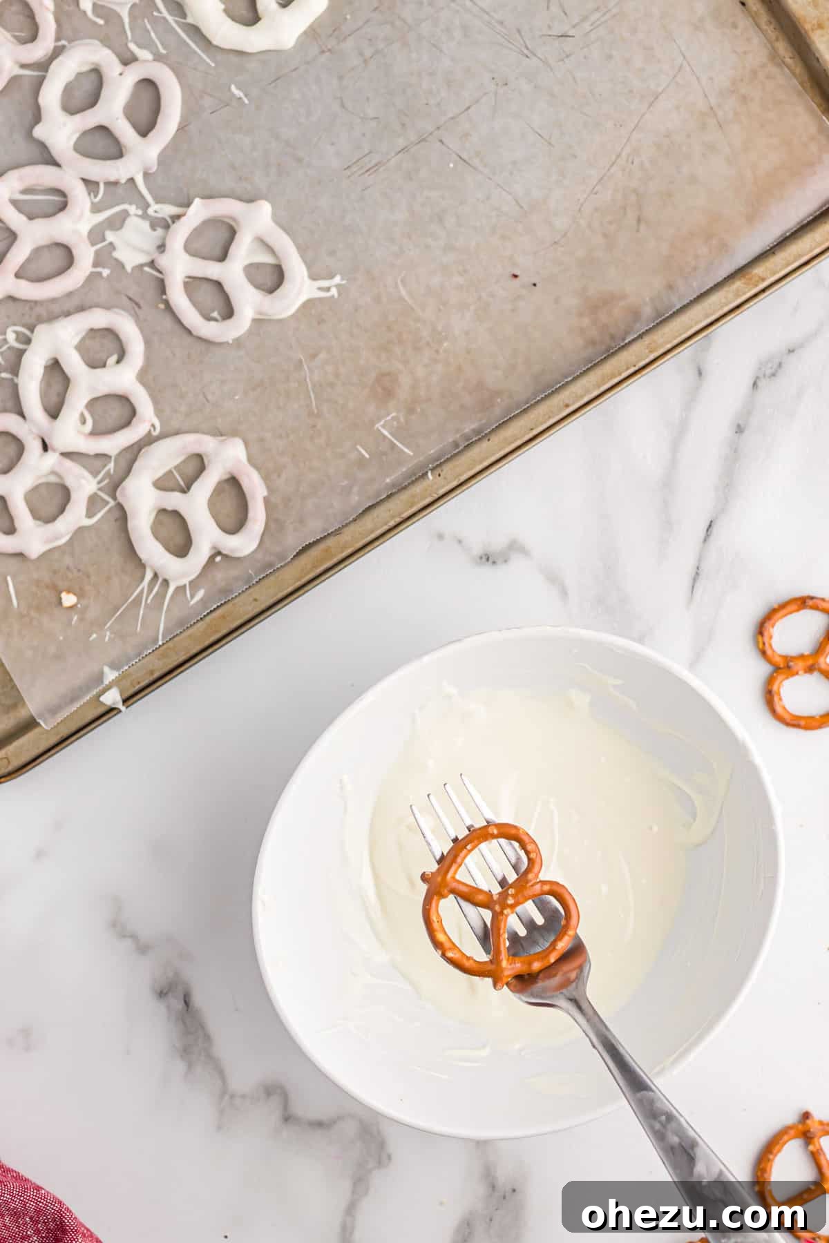 A hand holding a mini pretzel over a small bowl of melted white chocolate, demonstrating the dipping motion.