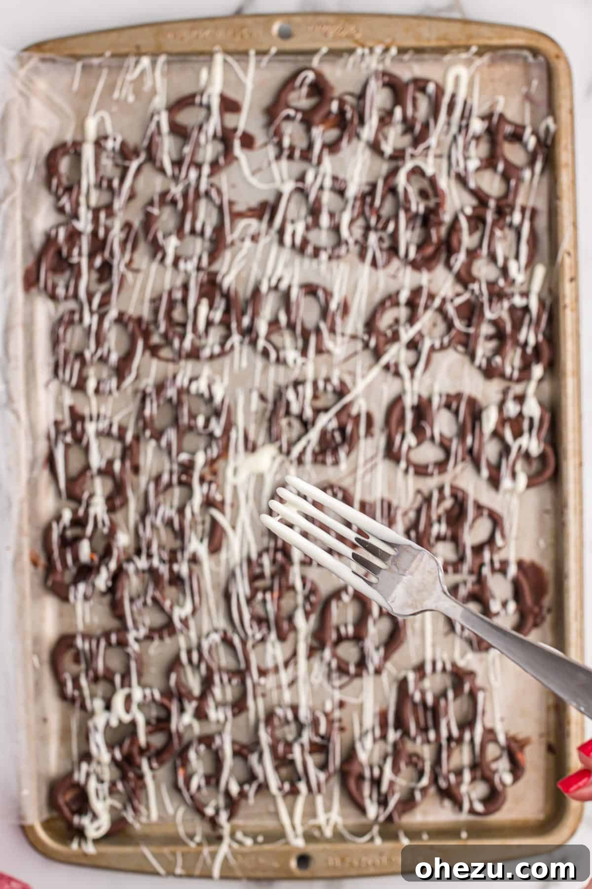 A close-up shot of white chocolate being drizzled over dark chocolate covered pretzels on a baking sheet, creating an attractive decorative pattern.