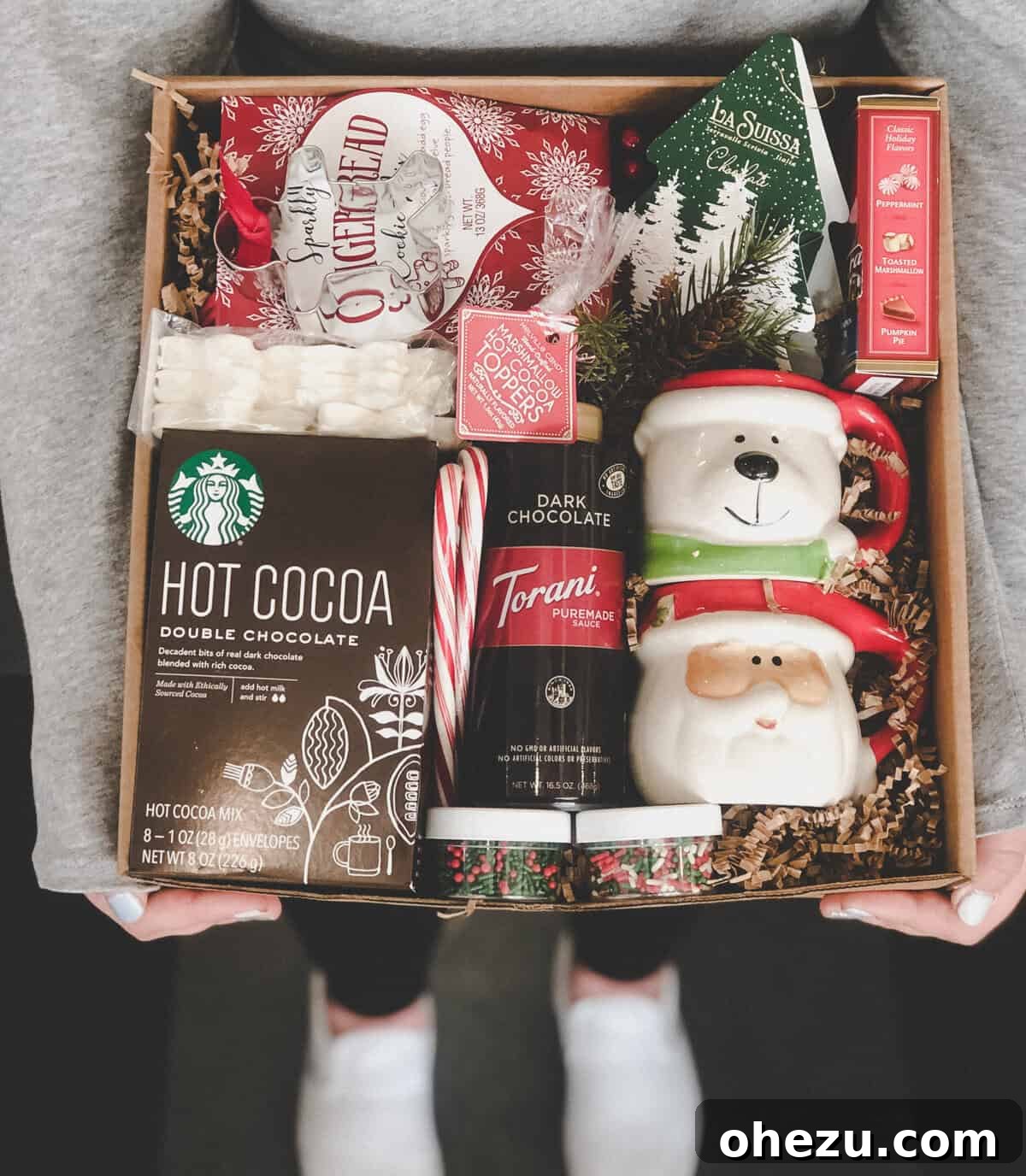 A curated gift basket featuring holiday mugs, cocoa mix, gingerbread, and various chocolates.