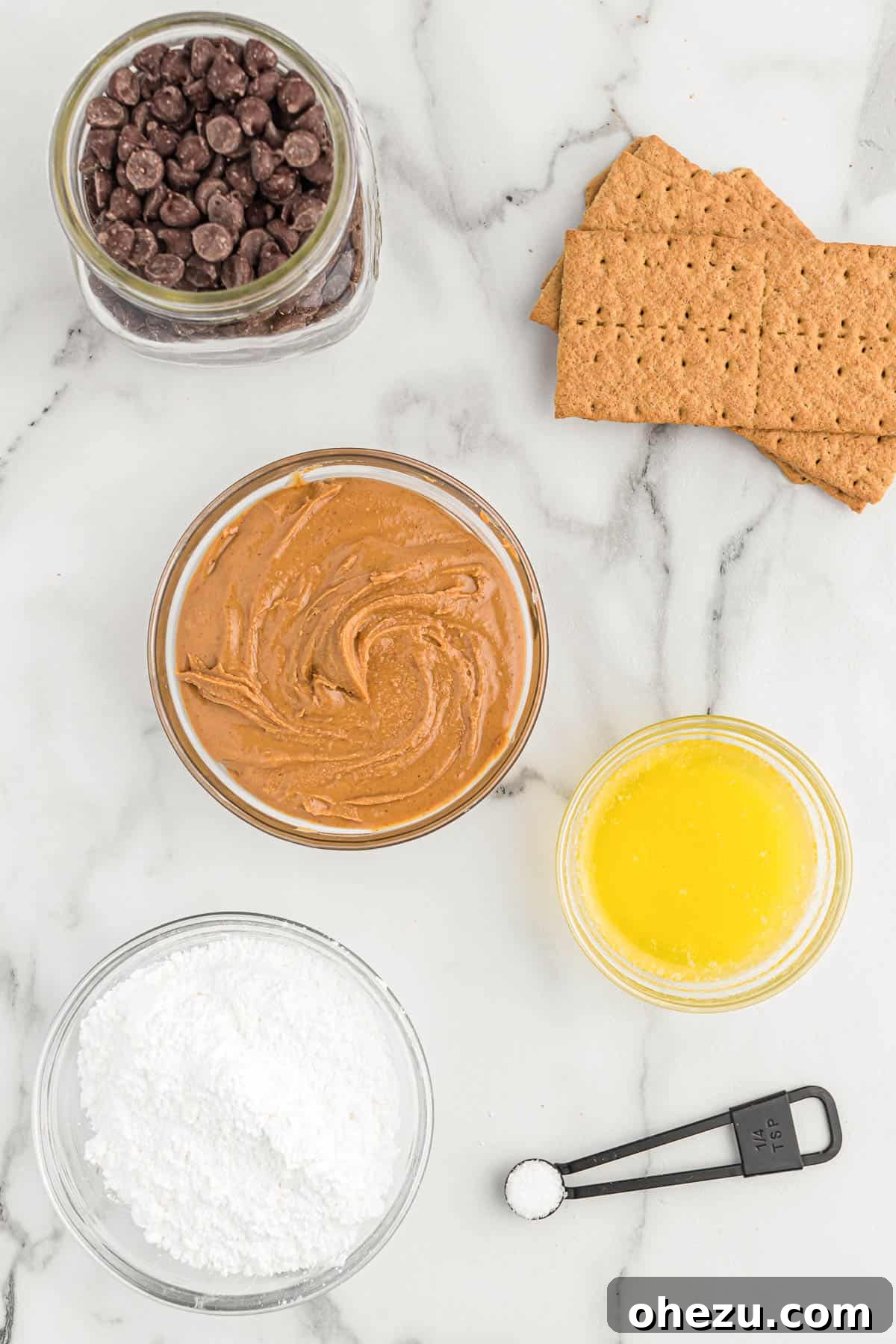 All six ingredients for peanut butter balls laid out on a white surface: peanut butter, butter, powdered sugar, graham crackers, salt, and chocolate chips