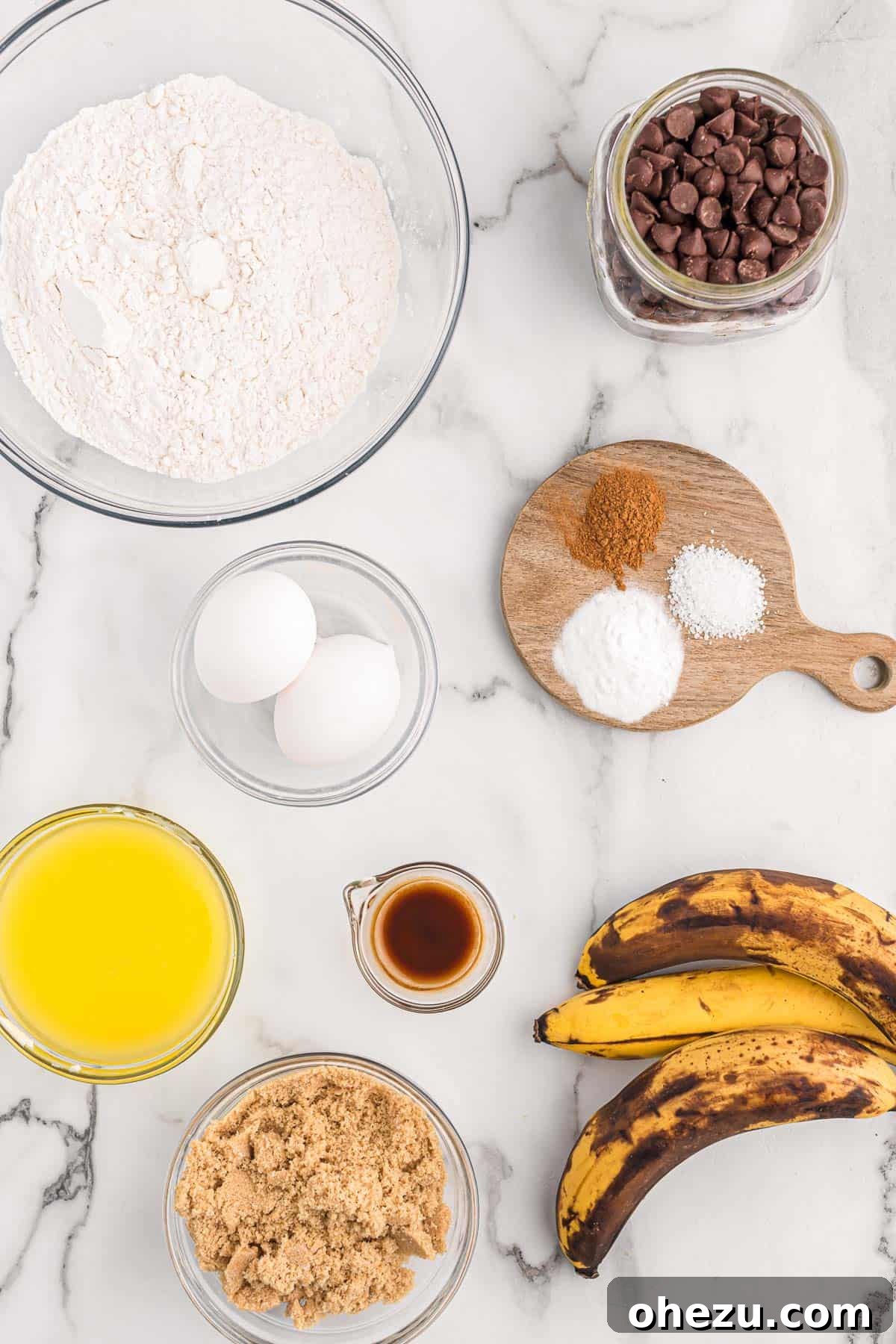 A vibrant collection of fresh ingredients carefully laid out on a clean kitchen counter, including several ripe, spotted bananas, a generous pile of semi-sweet chocolate chips, a stick of butter, eggs, bags of flour and brown sugar, all perfectly organized and ready for making the ultimate chocolate chip banana bread.