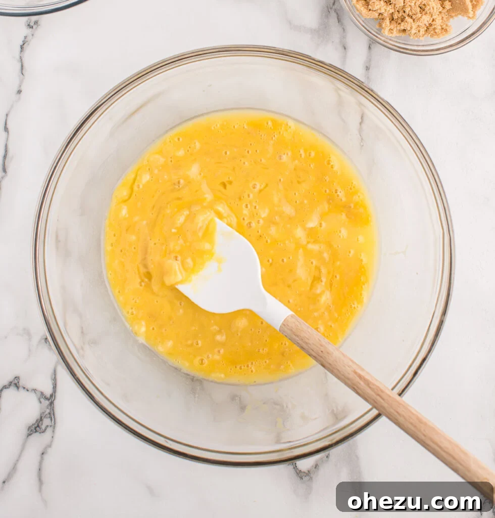 Wet ingredients for chocolate chip banana bread, including mashed ripe bananas, melted butter, eggs, and vanilla extract, being thoroughly mixed together in a large bowl.