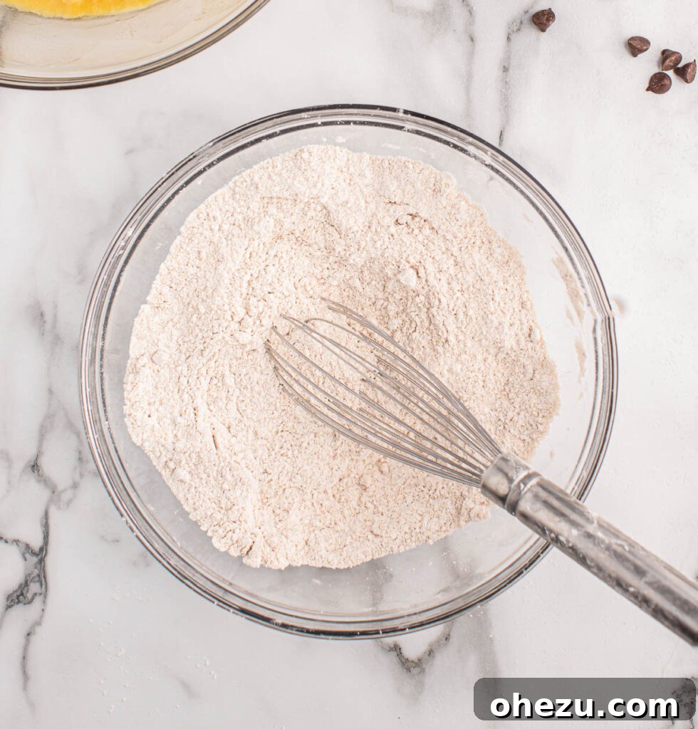 Dry ingredients for banana bread, including all-purpose flour, baking soda, salt, and cinnamon, being carefully whisked together in a separate, medium-sized bowl to ensure even distribution.