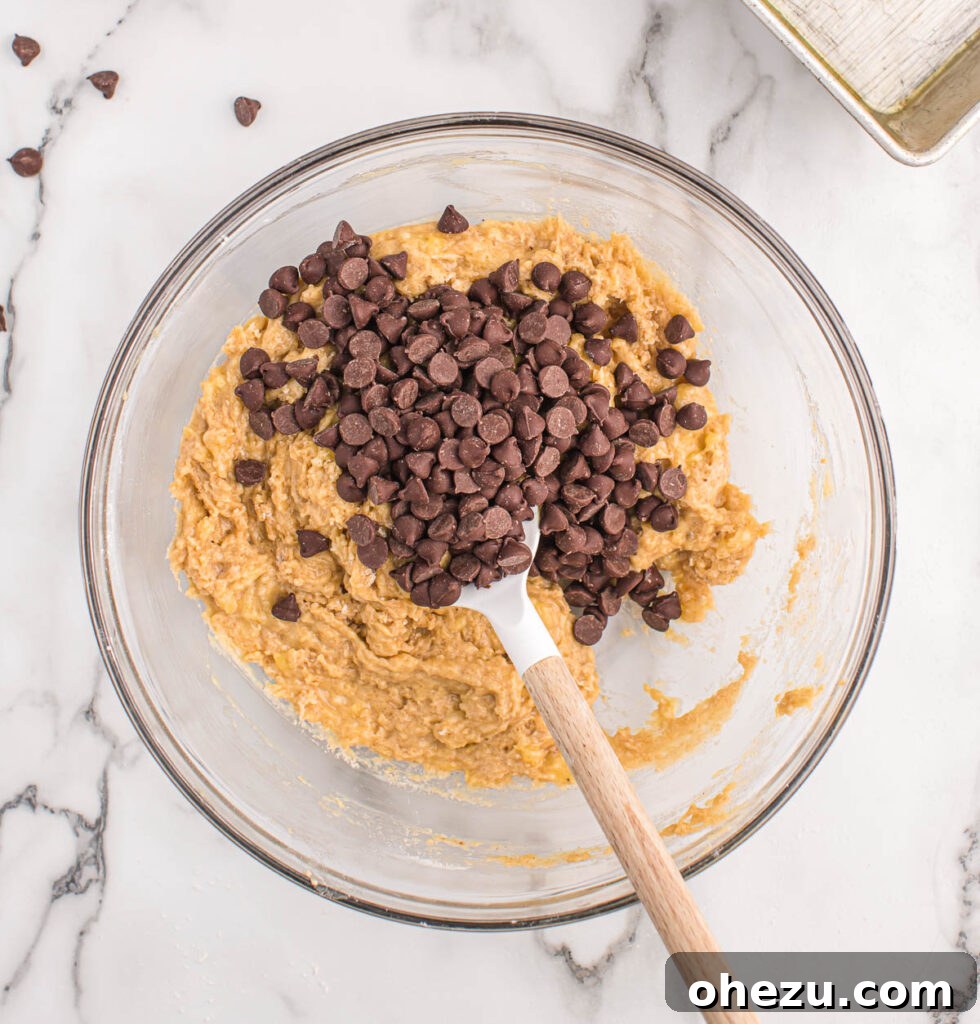 A wooden spatula gently stirring a generous amount of semi-sweet chocolate chips into the thick, rich banana bread batter in a mixing bowl, ensuring even distribution.