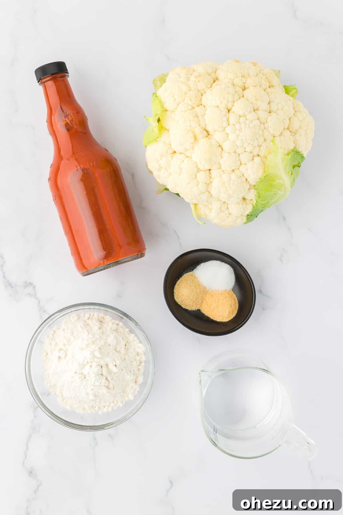 Individual ingredients laid out on a kitchen counter, including a head of cauliflower, a bowl of flour, separate small bowls of spices (salt, garlic powder, onion powder), and a bottle of buffalo sauce, all ready for the recipe.