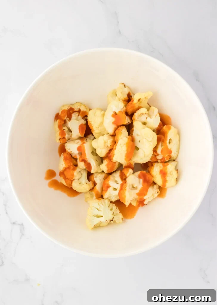 Crispy air-fried cauliflower being tossed with vibrant red buffalo sauce in a mixing bowl, ensuring full coverage.
