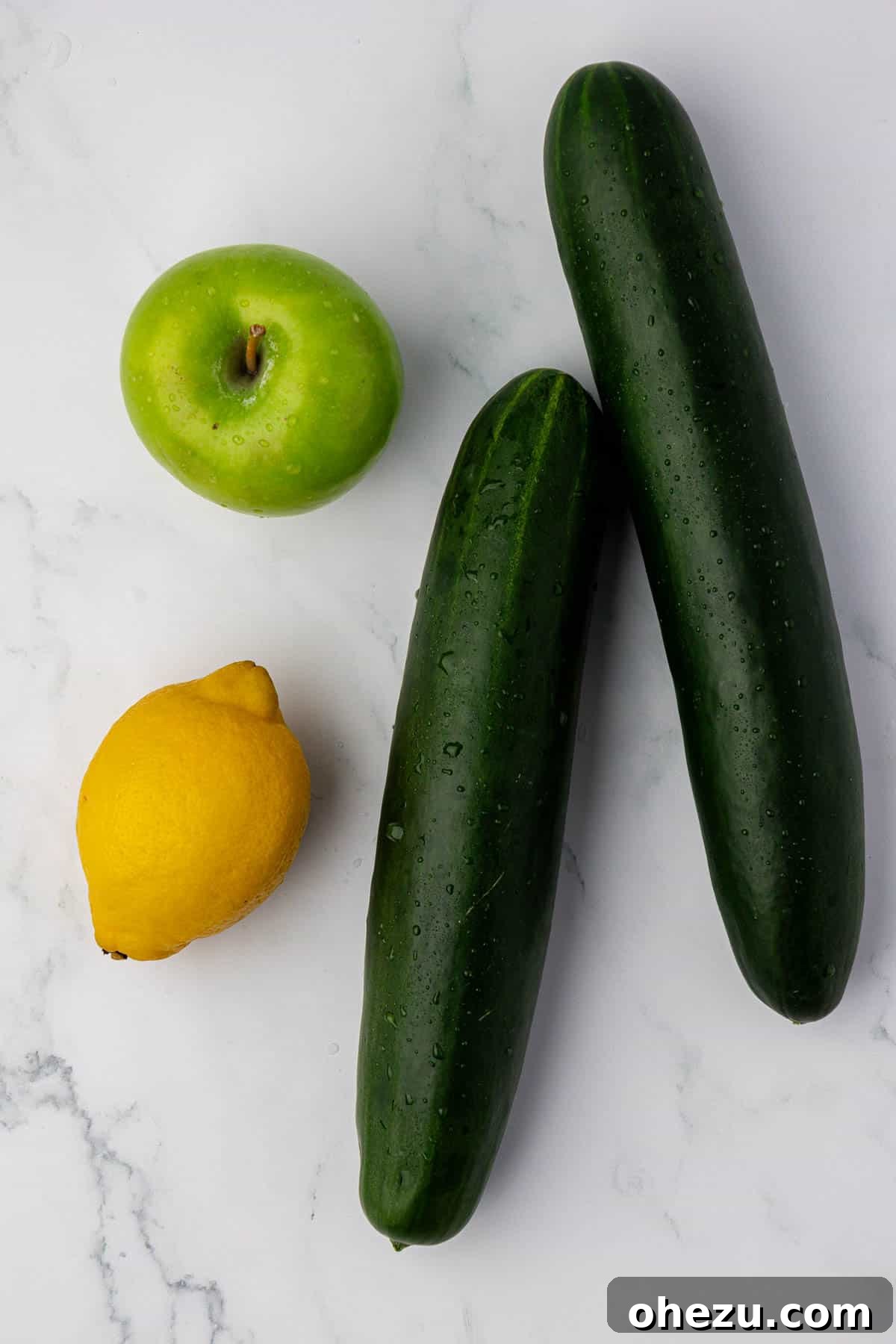 Two fresh cucumbers, a bright yellow lemon, and a vibrant green apple arranged on a surface, ready for juicing.