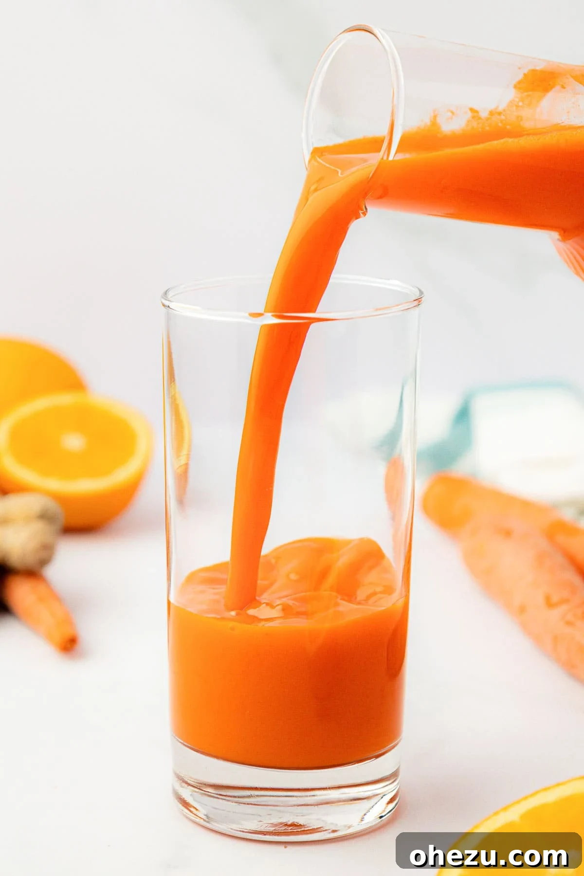 Pouring fresh, vibrant carrot juice from a glass pitcher into a tall serving glass with ice