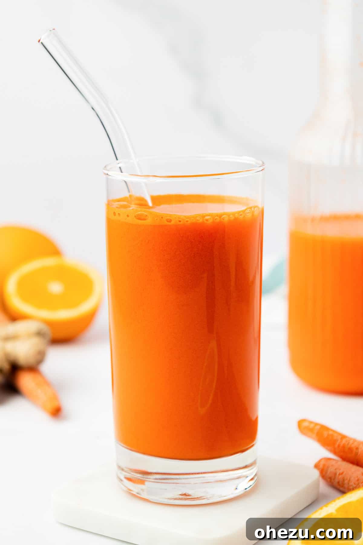 A tall glass of homemade carrot juice with a straw, accompanied by a fresh orange slice and a sprig of mint on a light background