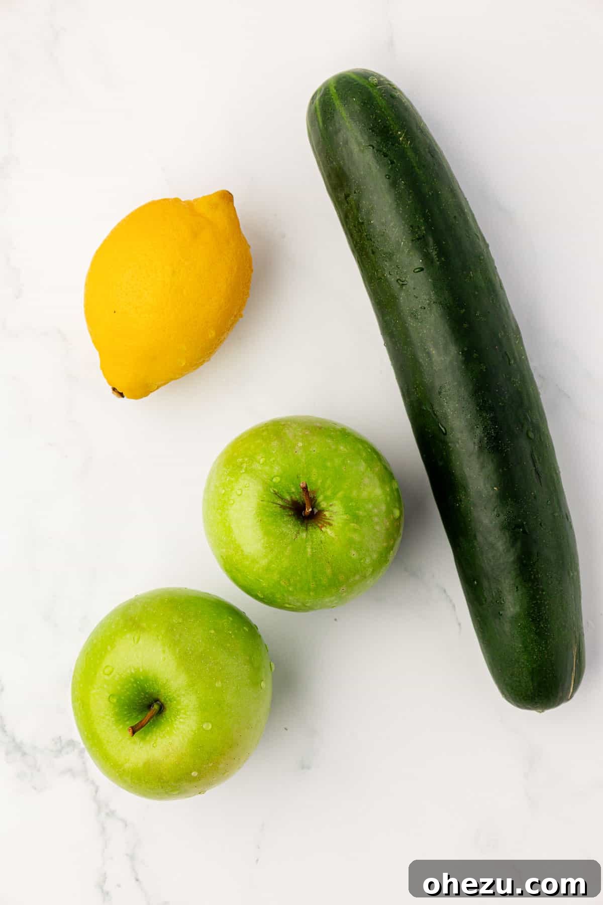 A simple still life of one lemon, two green apples, and a cucumber on a textured background