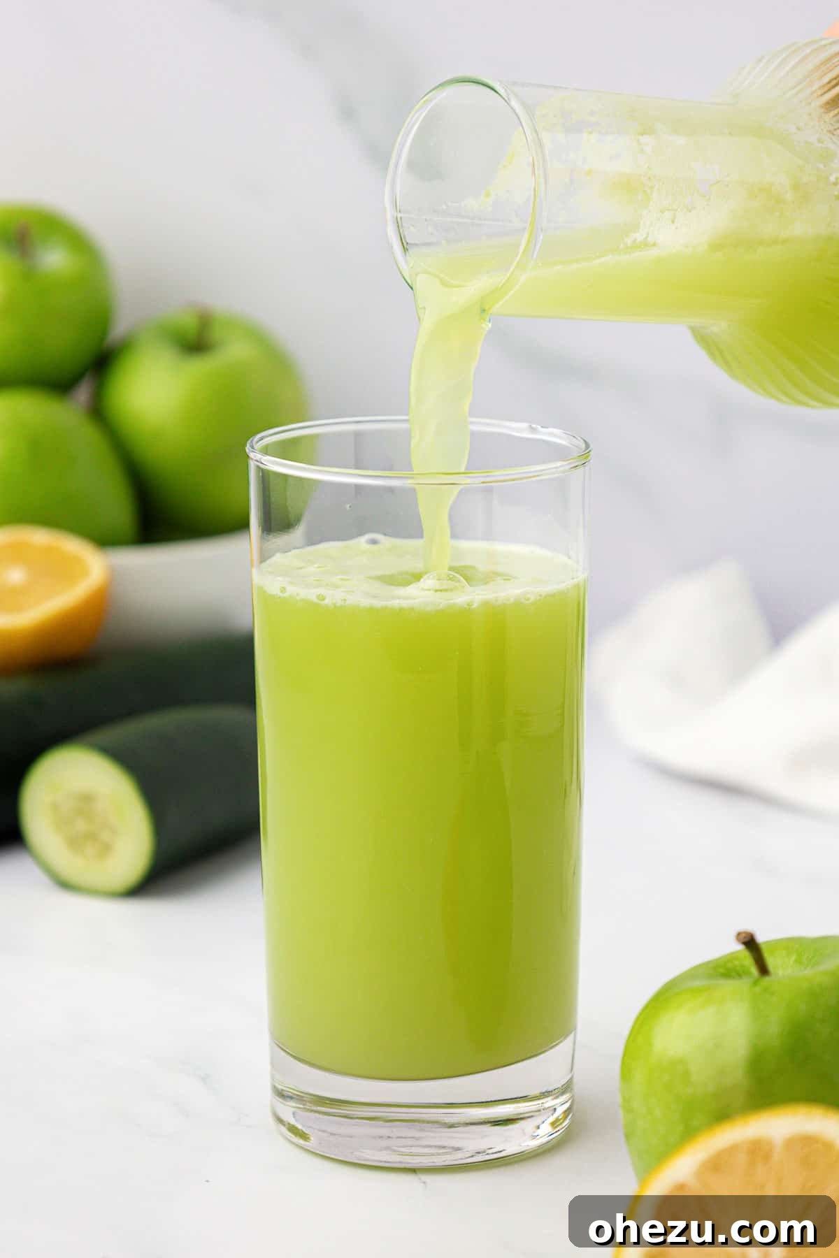 A hand pouring fresh green apple juice from a pitcher into a tall glass