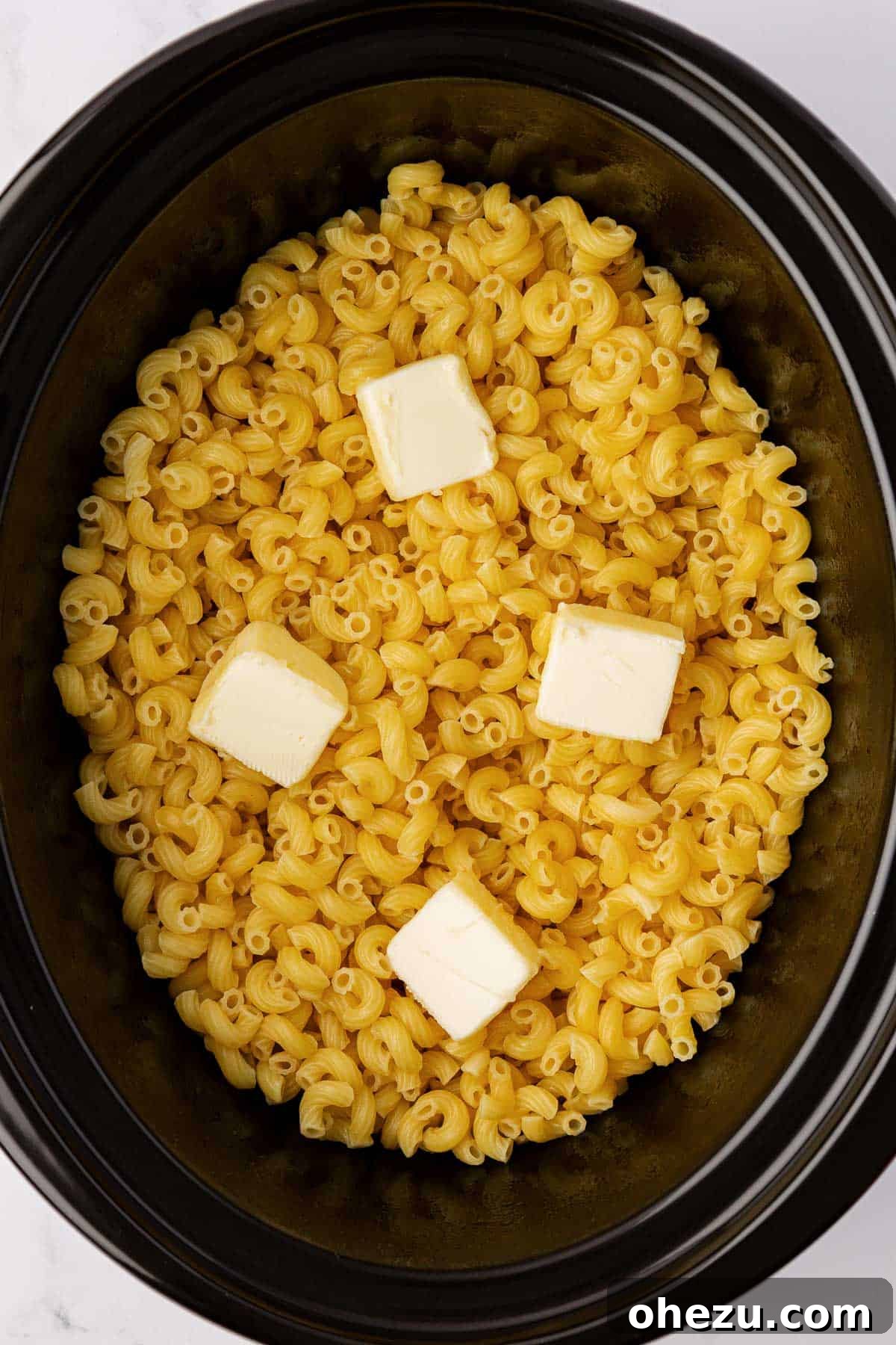 Creamy Slow Cooker Mac and Cheese 5 Cubed butter being stirred into partially cooked macaroni noodles in a crock pot, preparing for the cheese sauce.