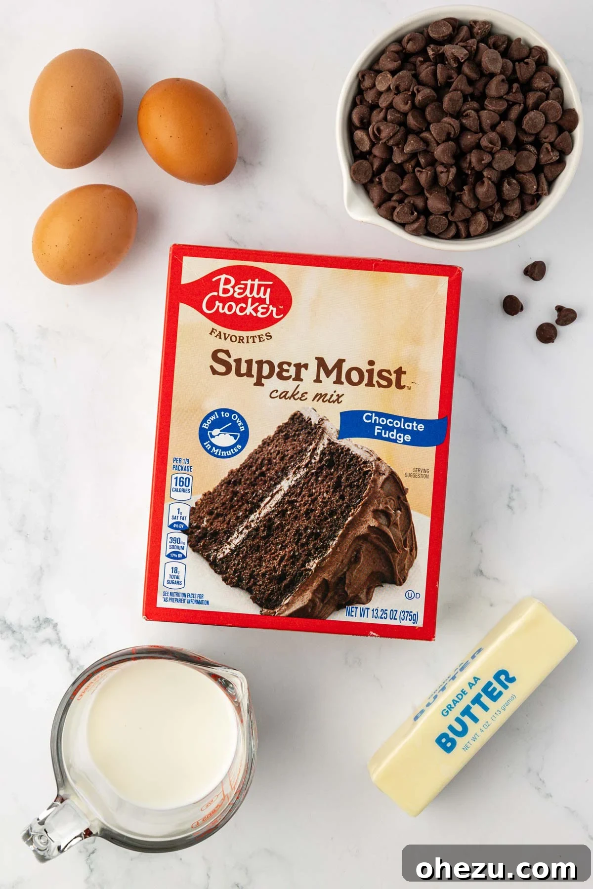 Individual ingredients laid out: a box of chocolate cake mix, fresh eggs, a bowl of chocolate chips, a glass of milk, and a stick of melted butter.