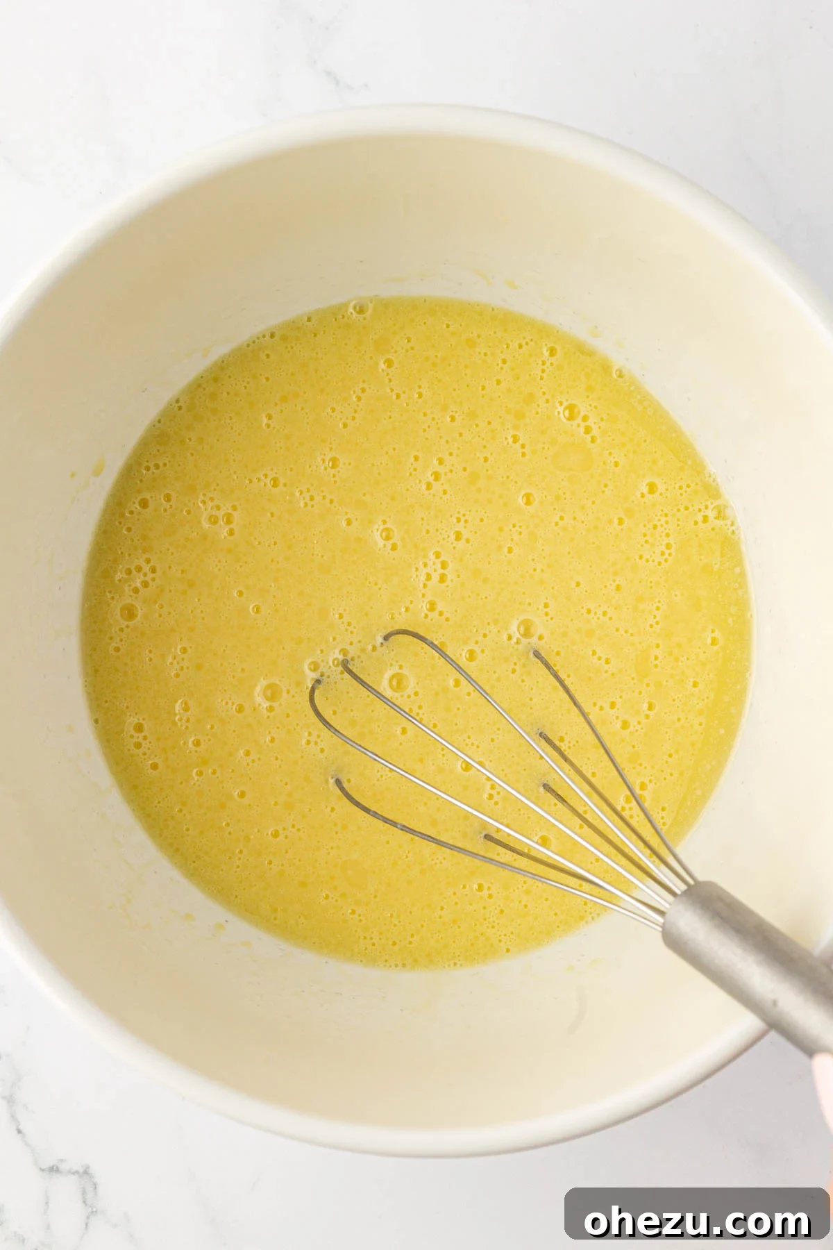 Melted butter and milk being whisked into beaten eggs in a mixing bowl, creating a uniform wet mixture.