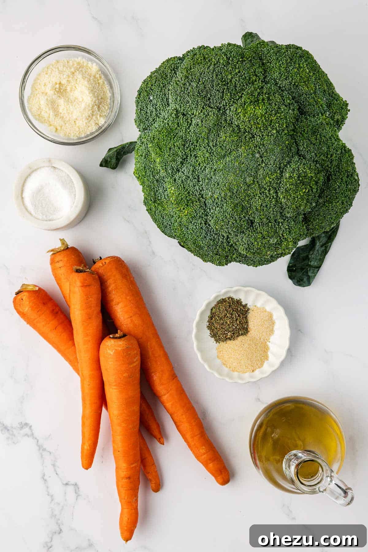 Ingredients for roasted broccoli and carrots laid out: a fresh head of broccoli, five carrots, grated parmesan cheese, olive oil, and a selection of spices.