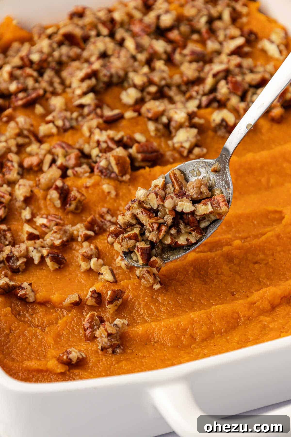 Hands sprinkling a generous layer of pecan crumble topping over the smooth sweet potato filling in a baking dish.