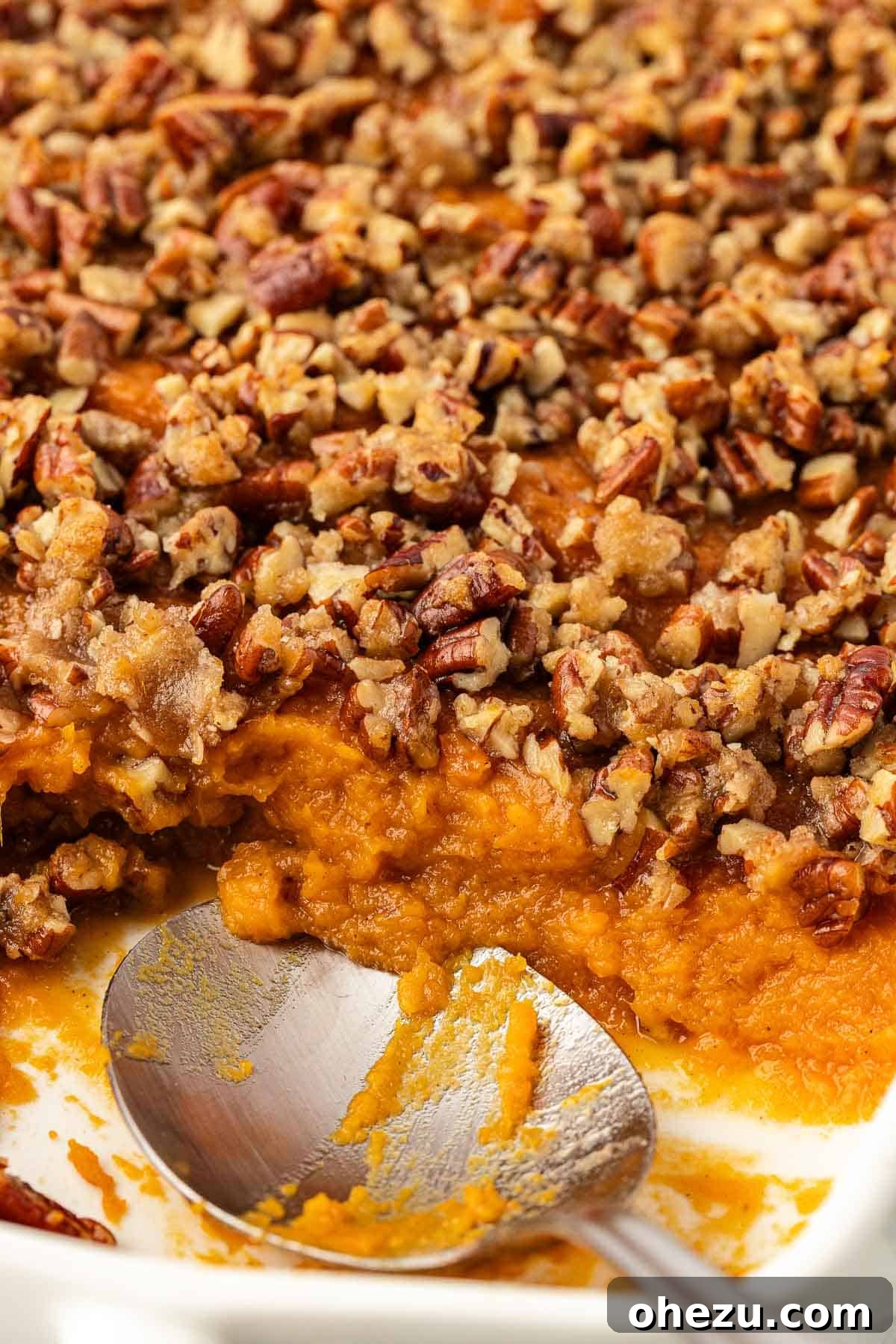 A sweet potato casserole with a scoop already taken out, revealing the creamy interior and the rich pecan topping.