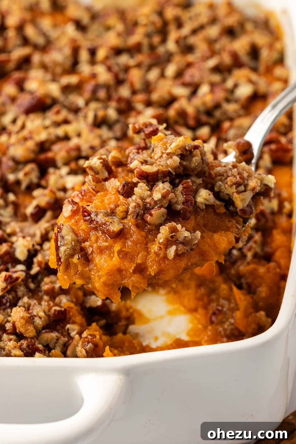 A serving spoon lifting a scoop of creamy sweet potato casserole with visible pecan topping from a baking dish.