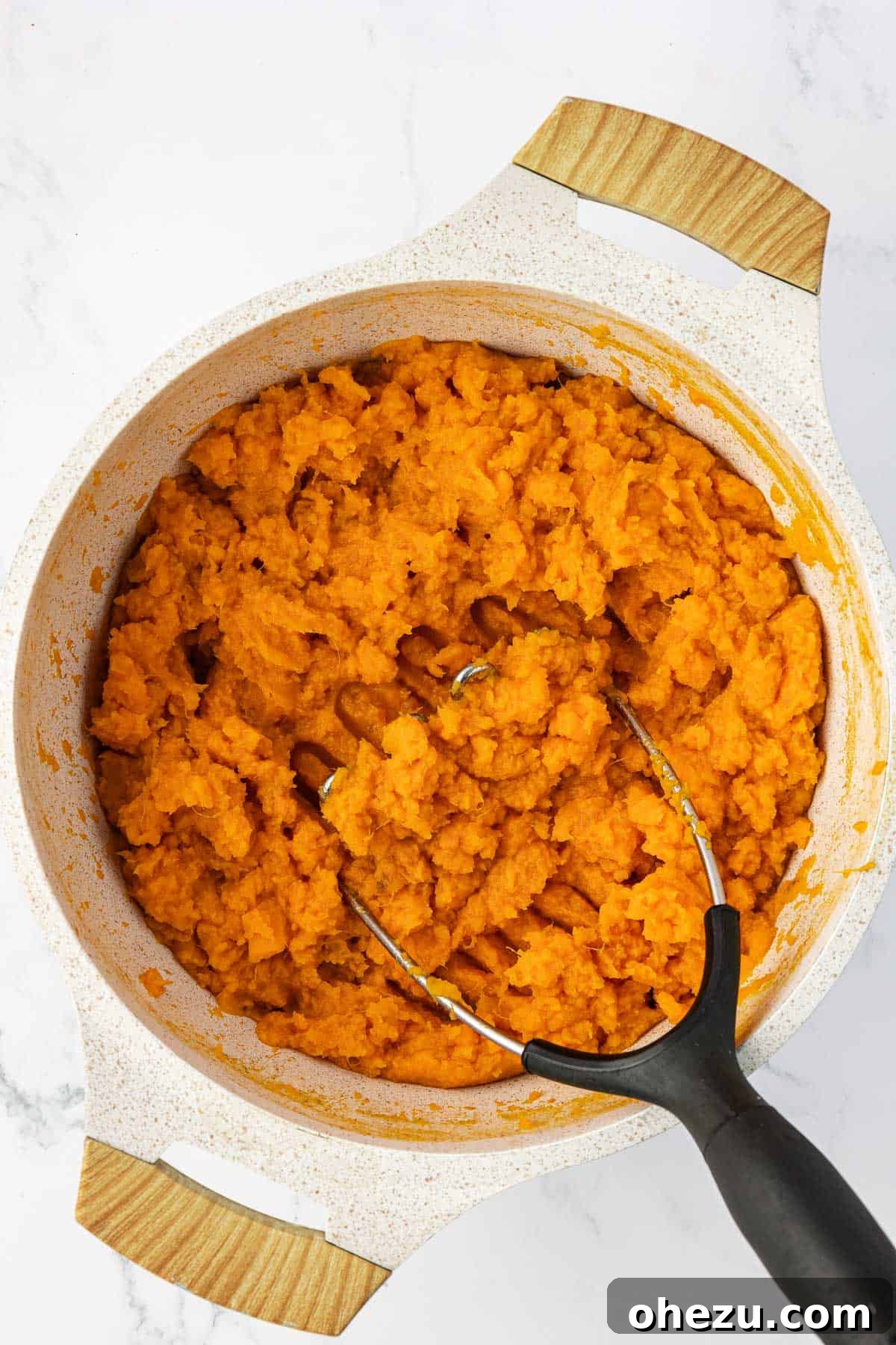 Hands mashing soft sweet potatoes in a large saucepan with a potato masher, creating a smooth, creamy base for the casserole.