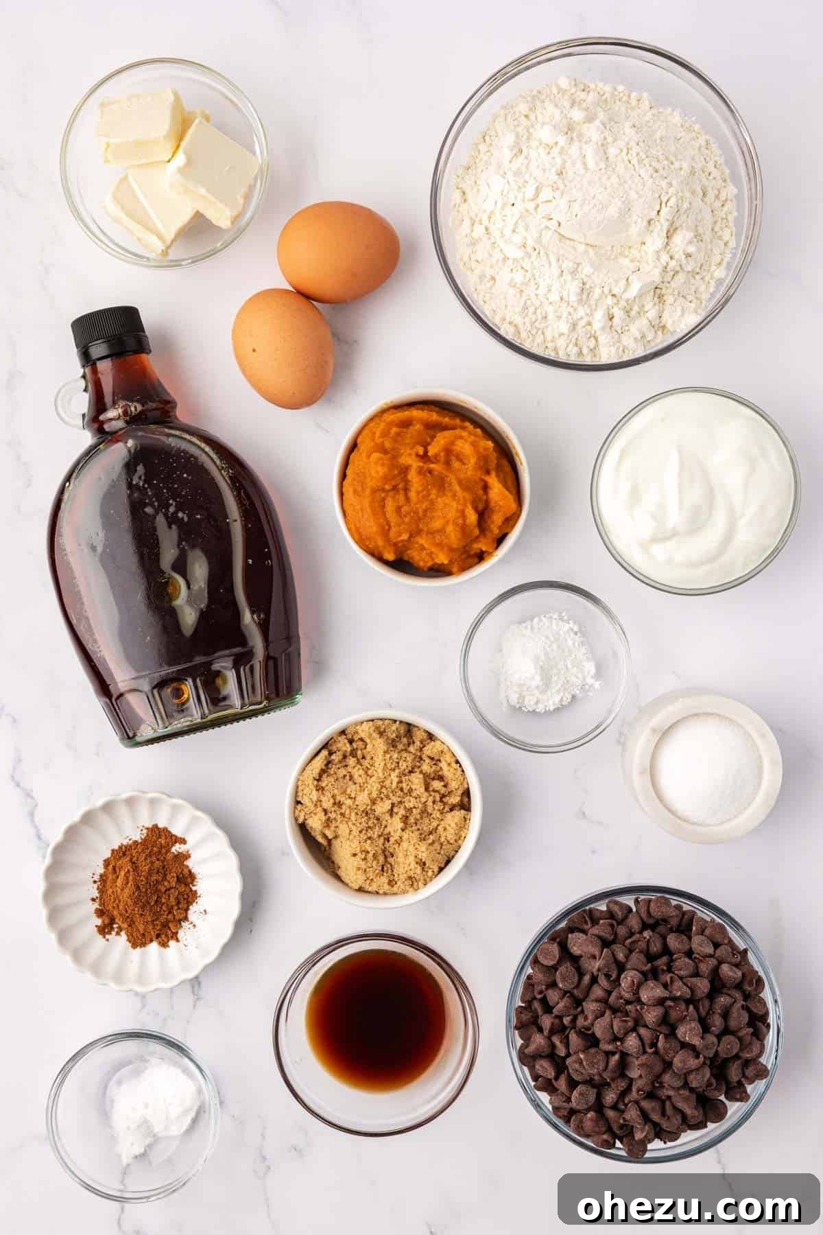 All the essential ingredients for making pumpkin chocolate chip muffins laid out on a kitchen counter: Greek yogurt, pumpkin puree, butter, eggs, vanilla, spices, maple syrup, brown sugar, flour, and chocolate chips.