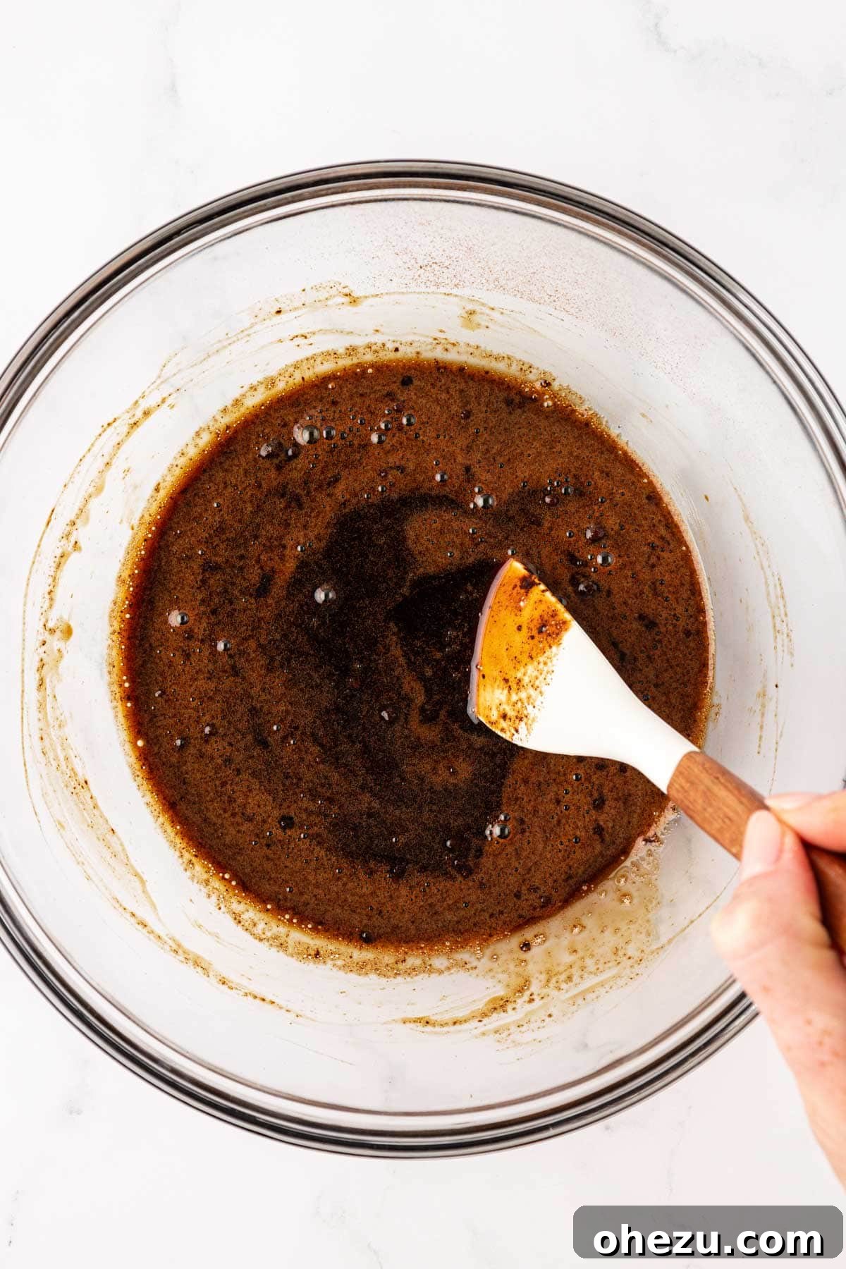 A rubber spatula mixing instant espresso powder into maple syrup until fully incorporated.