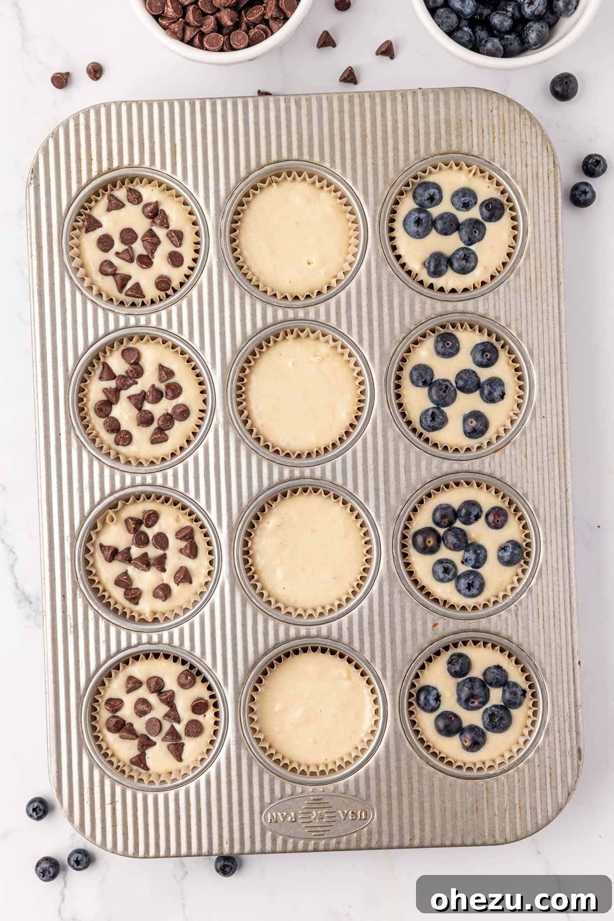 Pancake muffin batter poured into a muffin tin lined with parchment paper cups, topped with blueberries and chocolate chips.