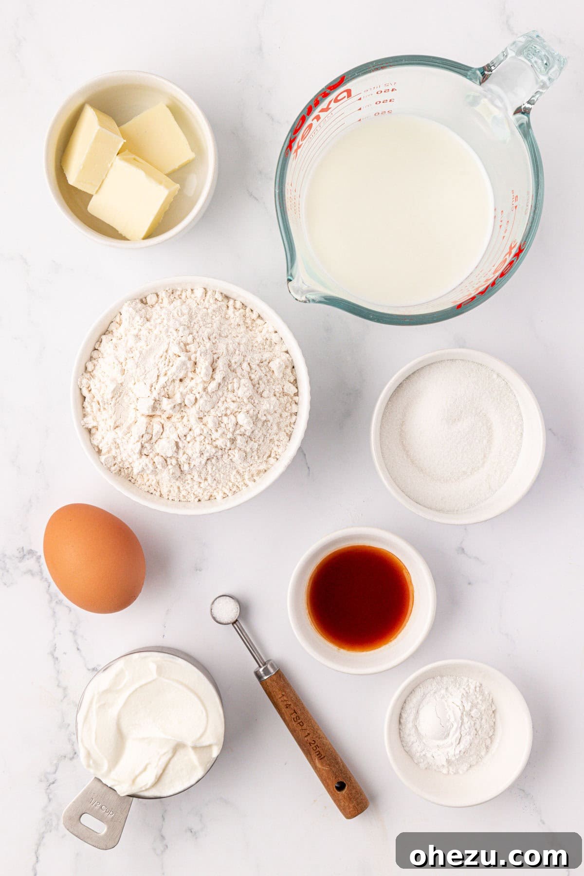 A selection of ingredients laid out on a kitchen counter, including flour, sugar, milk, Greek yogurt, butter, egg, and vanilla extract, ready for making pancake muffins.