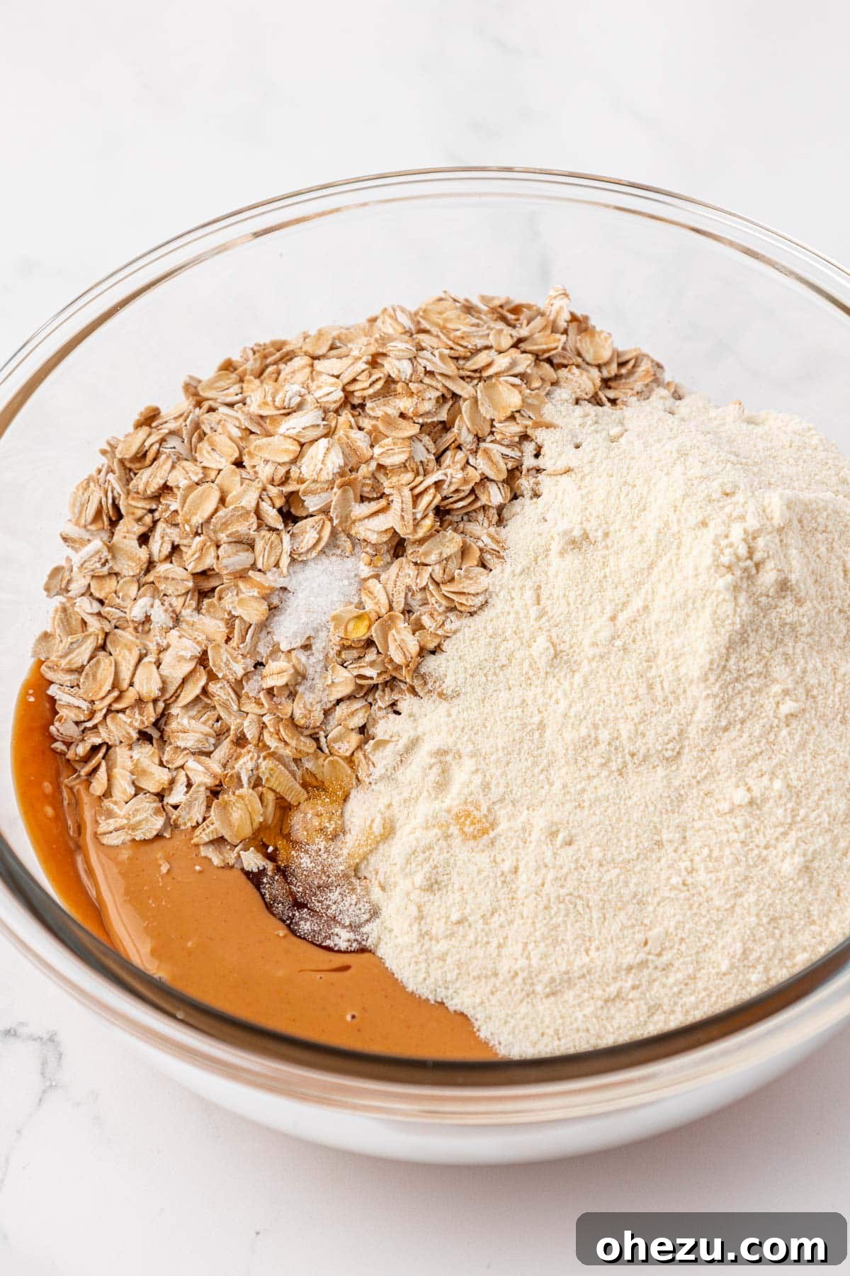 All the dry and liquid ingredients, including honey, vanilla protein powder, rolled oats, and salt, are being added to a large mixing bowl containing melted peanut butter.