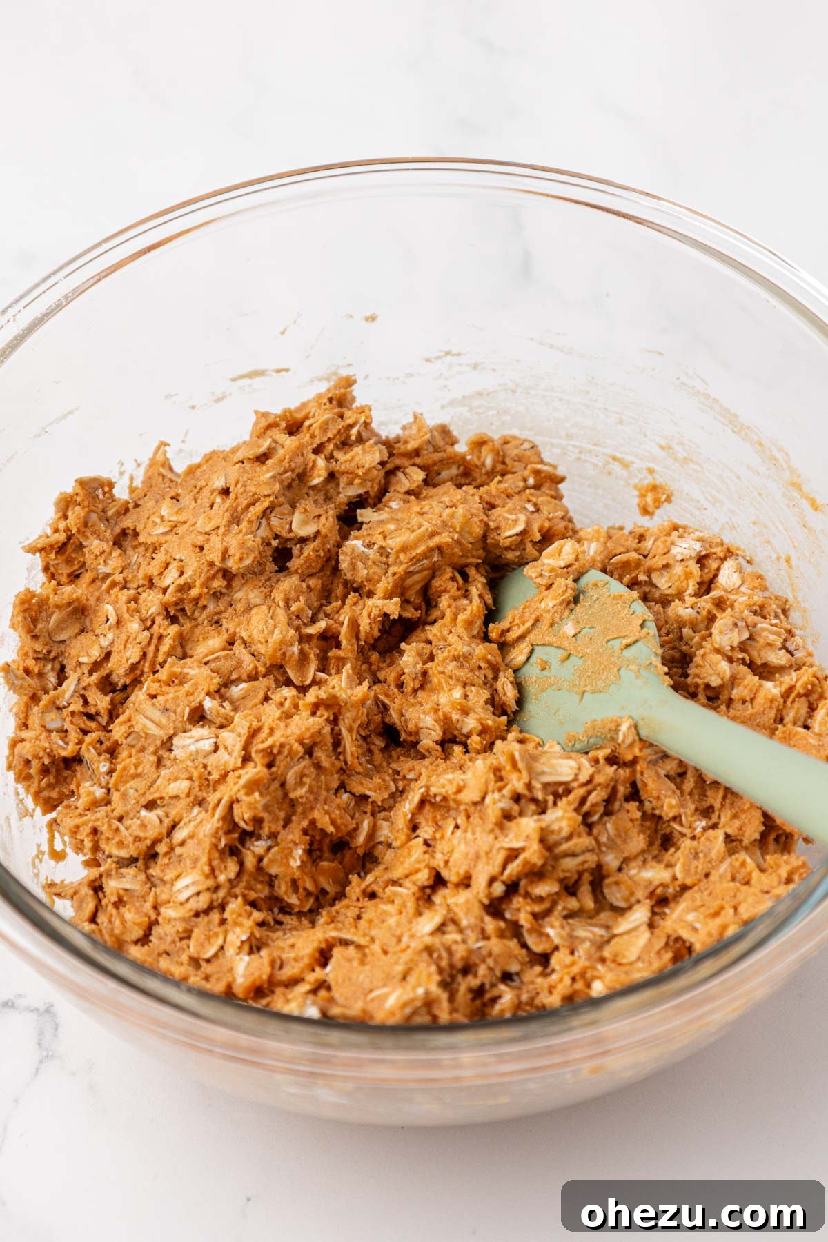 A rubber spatula is actively stirring the combined protein granola bar mixture in a bowl, showcasing the thick, uniform consistency of the batter before any chocolate chips are incorporated.
