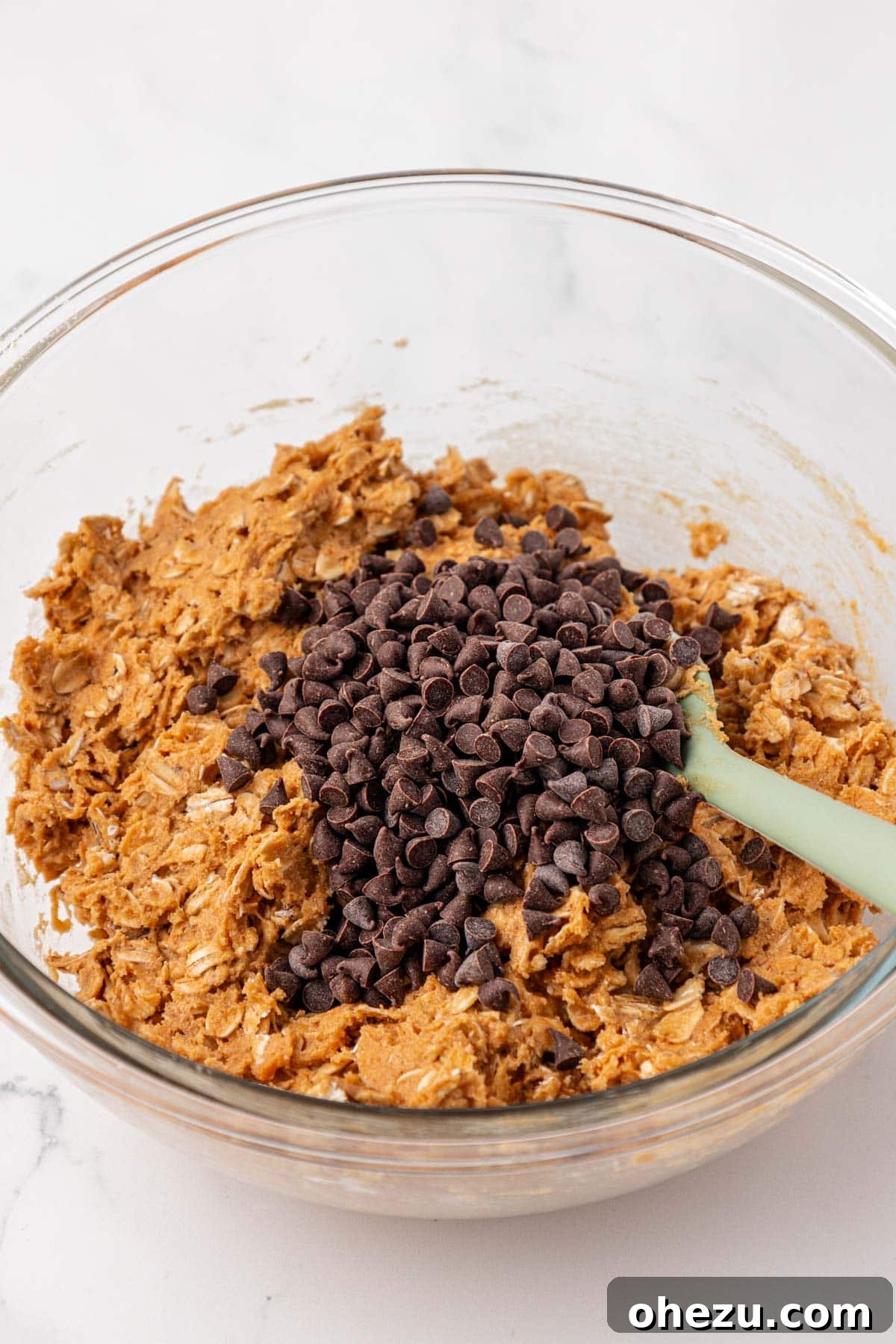 Mini chocolate chips being carefully added to the cooled and mixed protein granola bar batter in a large mixing bowl.