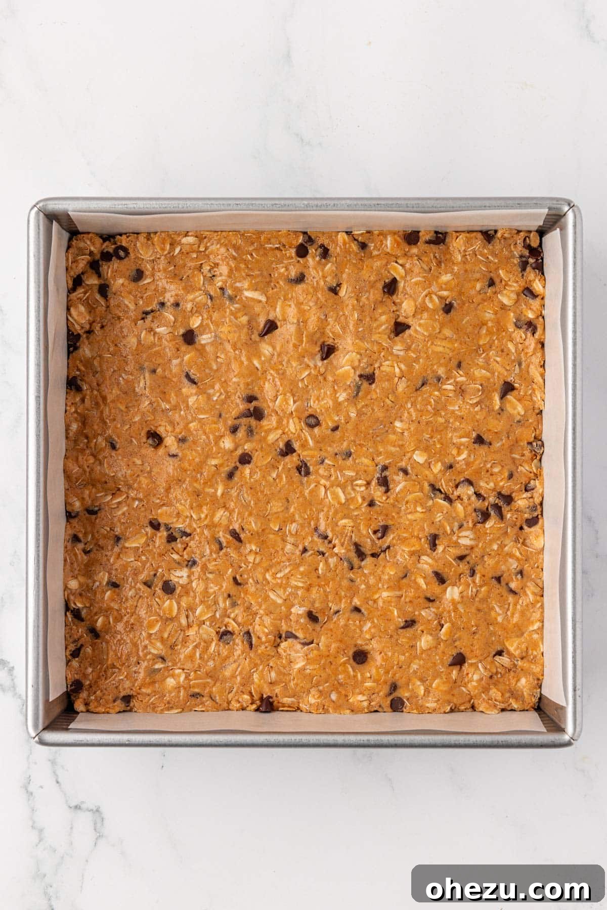 Protein granola bar batter expertly pressed into an 8x8 inch baking dish lined with parchment paper, prepared for refrigeration.