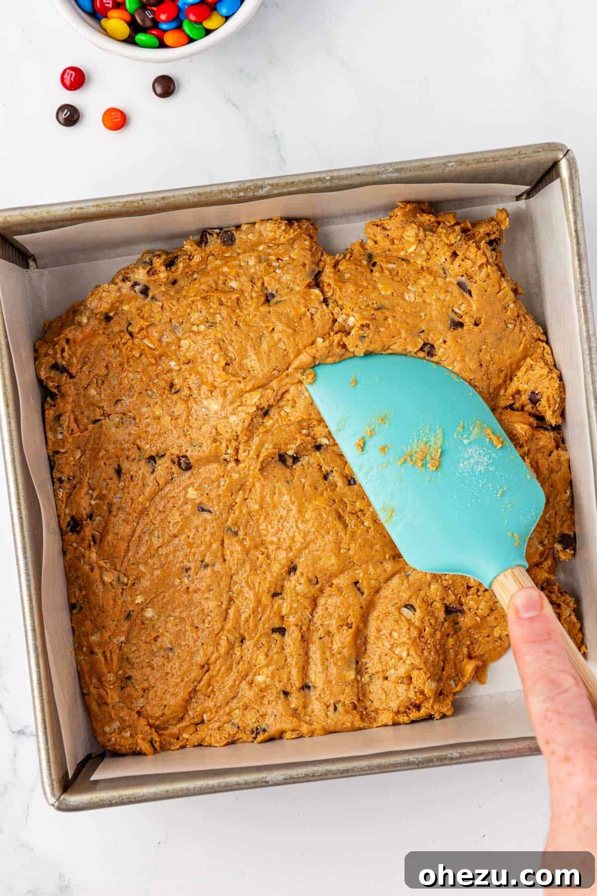 Rubber spatula pressing monster cookie protein bar batter into a parchment-lined pan